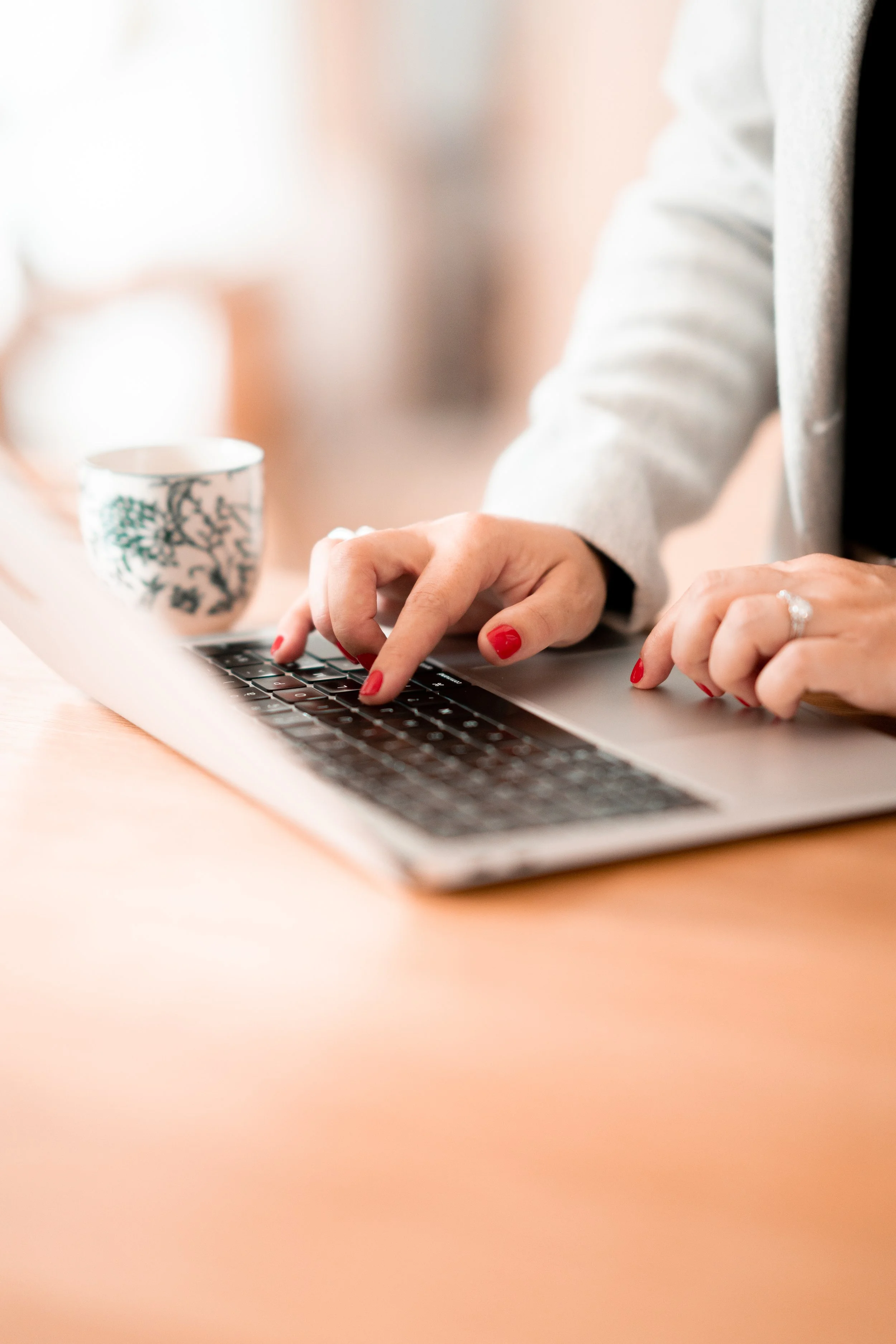 Une personne tape sur le clavier d'un ordinateur portable. La personne porte du vernis à ongles rouge et une bague. Il y a une tasse décorative blanche et verte à côté de l'ordinateur.