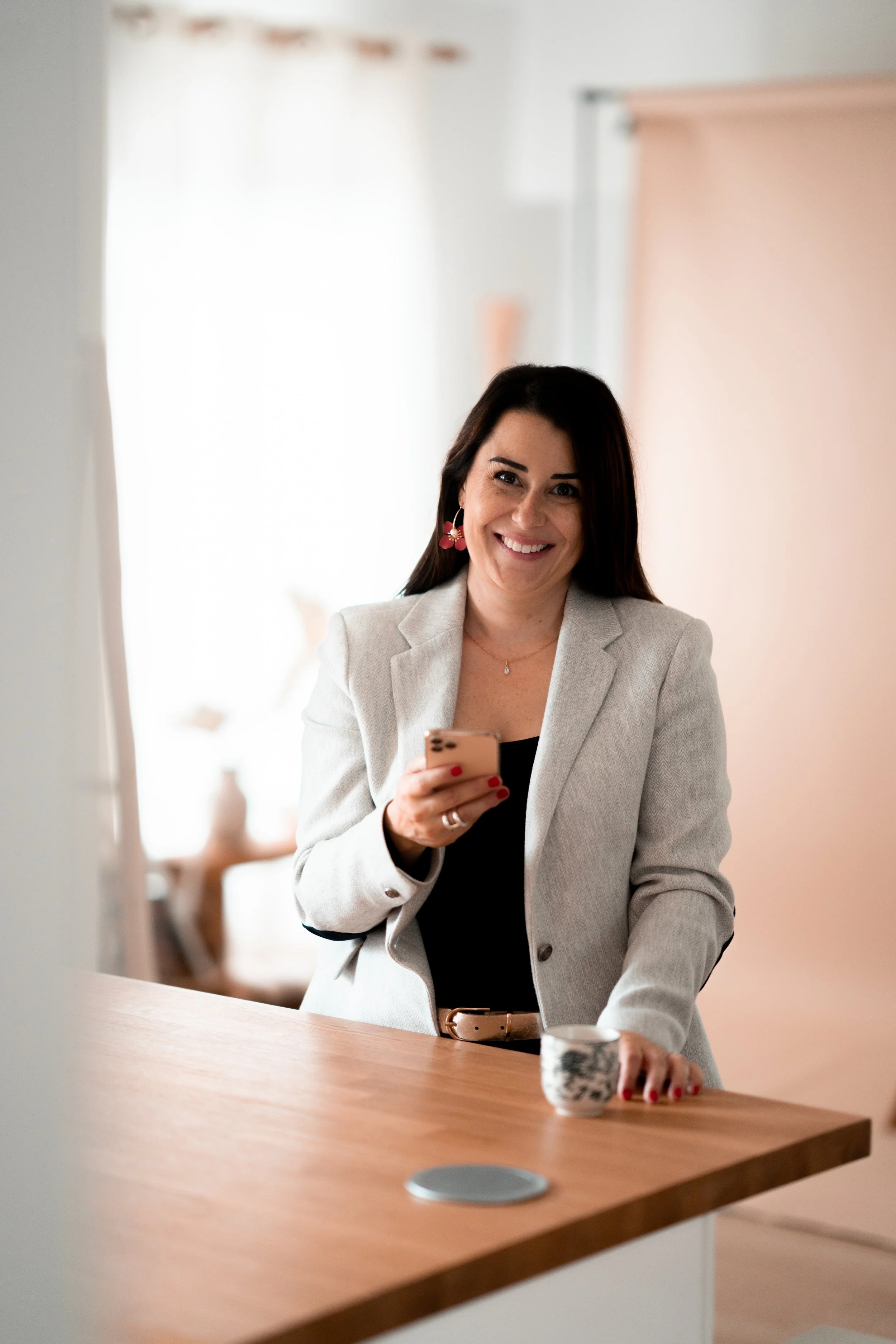 Une femme souriante en blazer gris et boucle d'oreilles colorées, tenant un smartphone, assise à une table en bois avec une tasse à portée de main dans un intérieur lumineux.