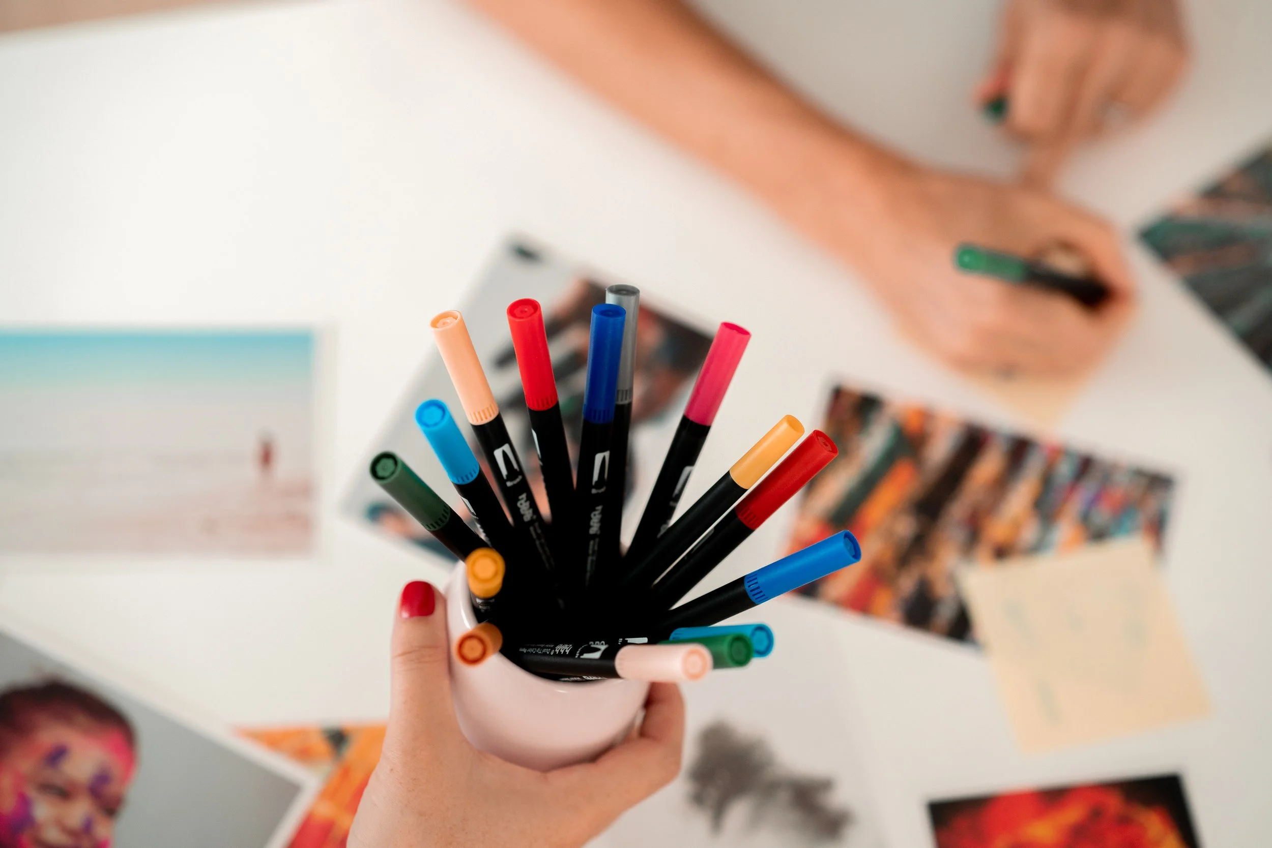 Une main tenant un pot avec plusieurs marqueurs de différentes couleurs. À l'arrière-plan, une personne écrit quelque chose sur une feuille lors d'un atelier de créativité.