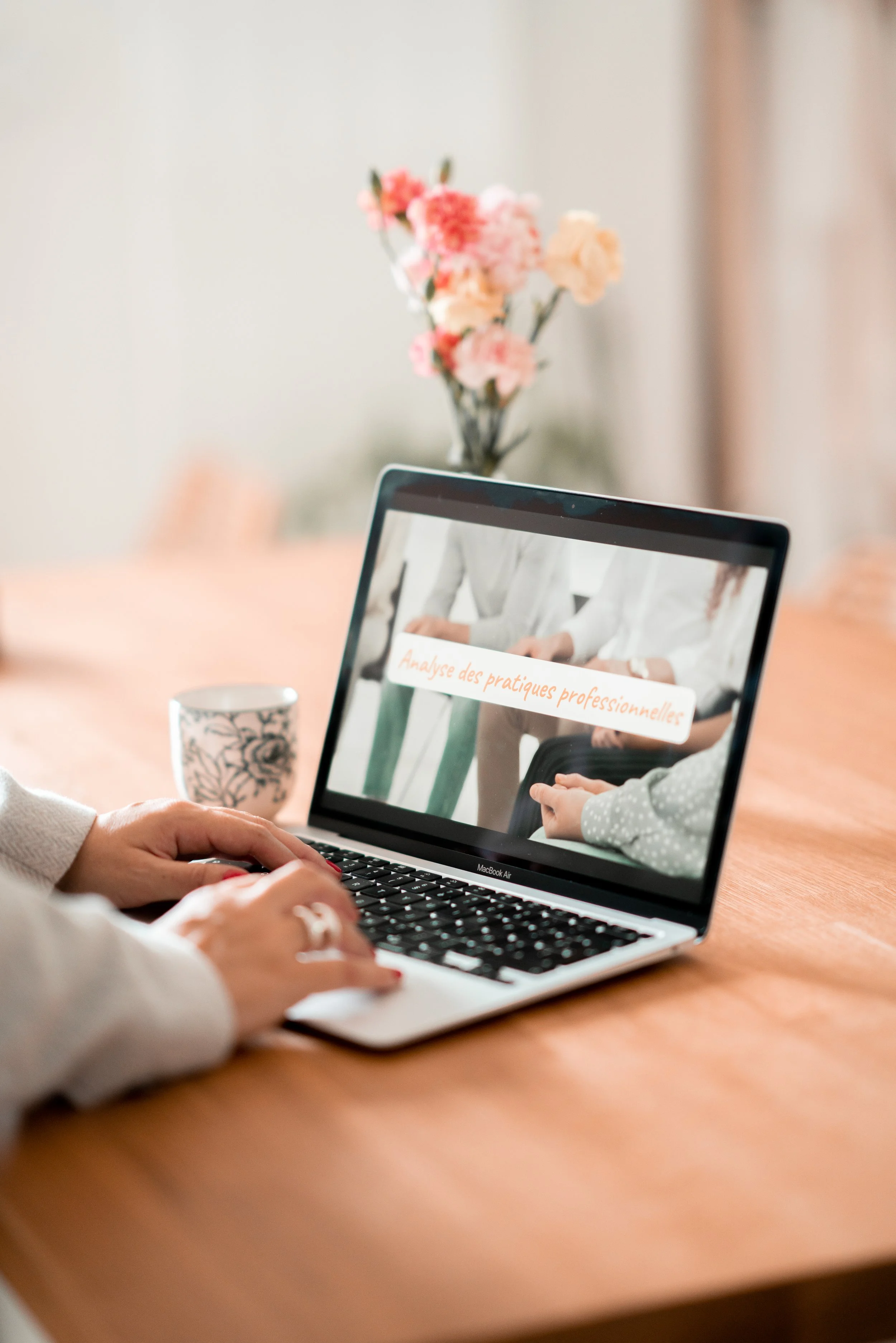 Une personne tape sur un ordinateur portable sur une table en bois, avec un mug à côté et un vase de fleurs en arrière-plan.