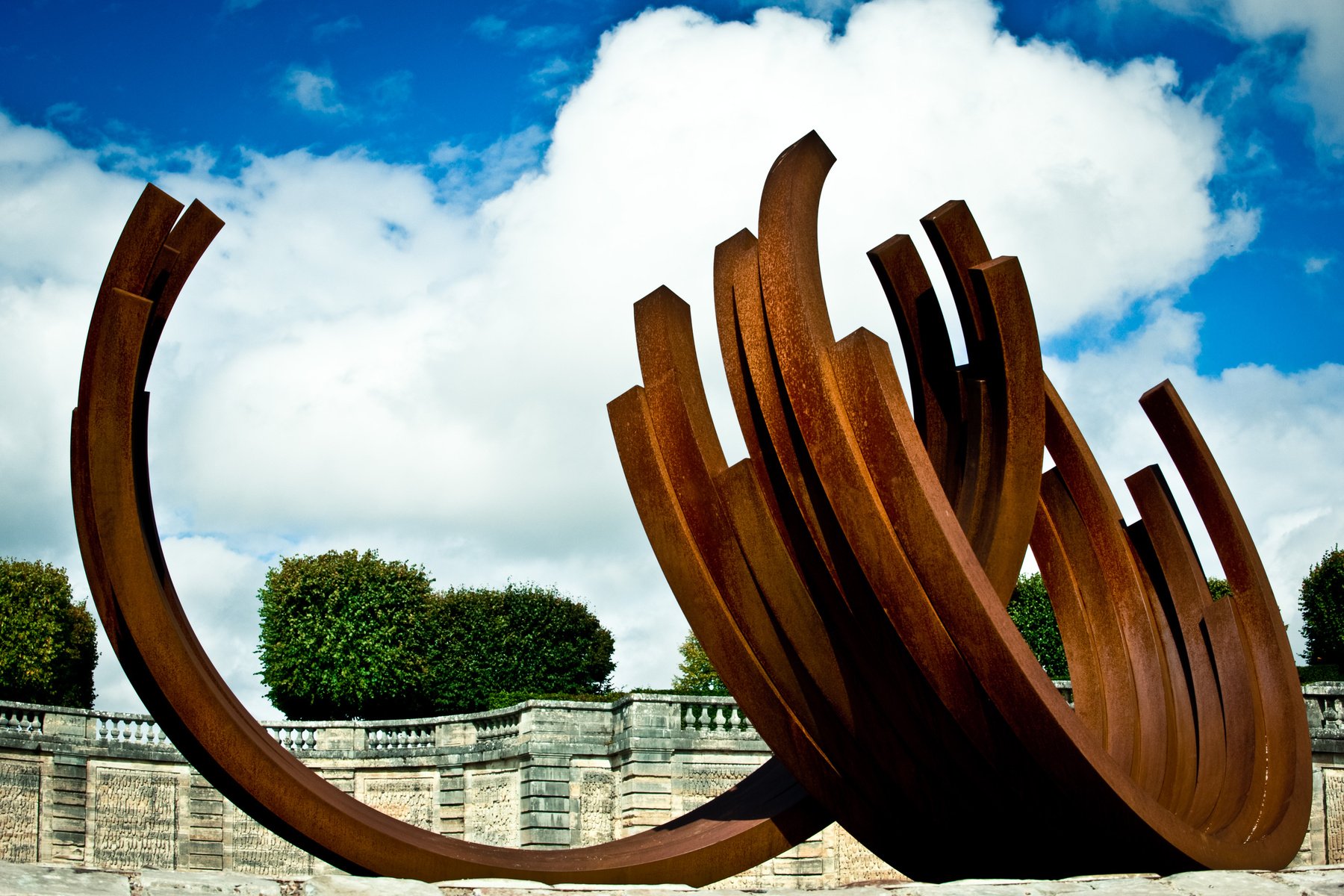 Large rust-colored metal sculpture with curved, elongated shapes outdoors against a partly cloudy sky and green trees.