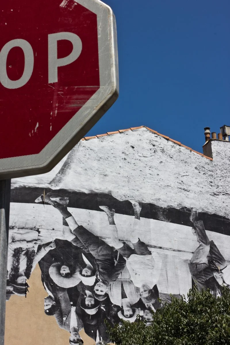 A red stop sign in the foreground with a mural of people hanging upside down on a wall in the background, against a blue sky.