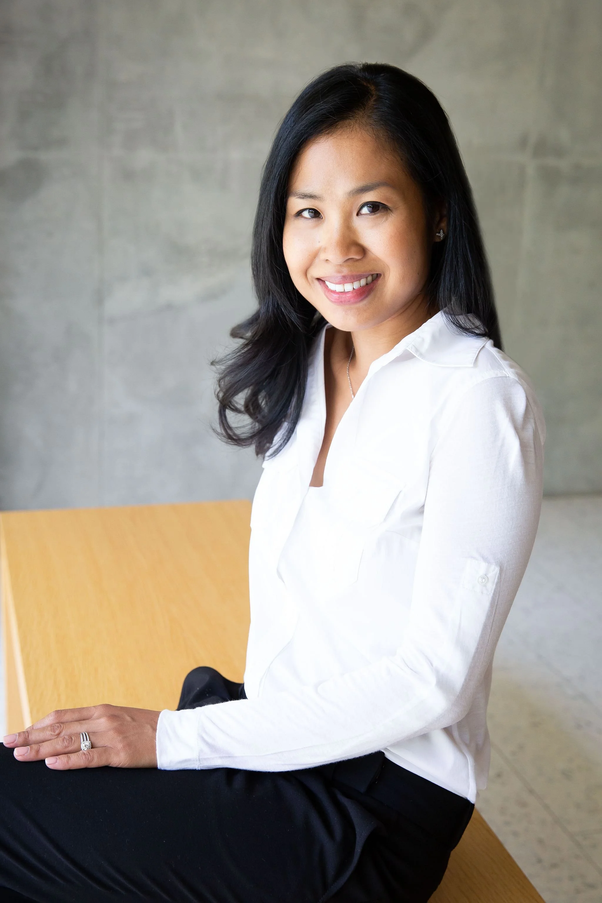 A woman with shoulder-length black hair, smiling, wearing a white blouse and black pants, sitting on a wooden bench against a concrete wall.