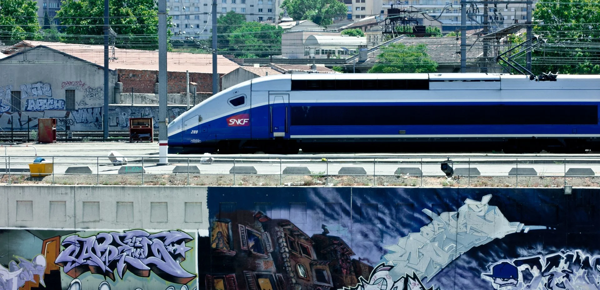 A modern high-speed train with a sleek white and blue design on railway tracks, with graffiti and city buildings in the background.