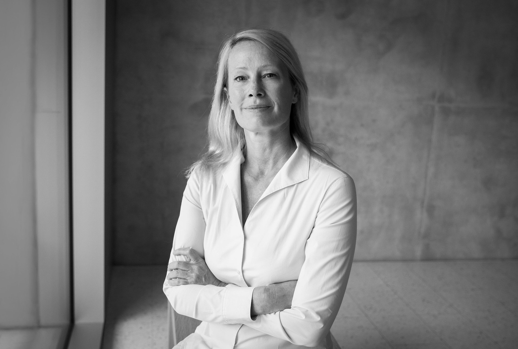 A middle-aged woman with blonde hair in a white shirt sitting with her arms crossed, looking at the camera, in front of a plain wall.
