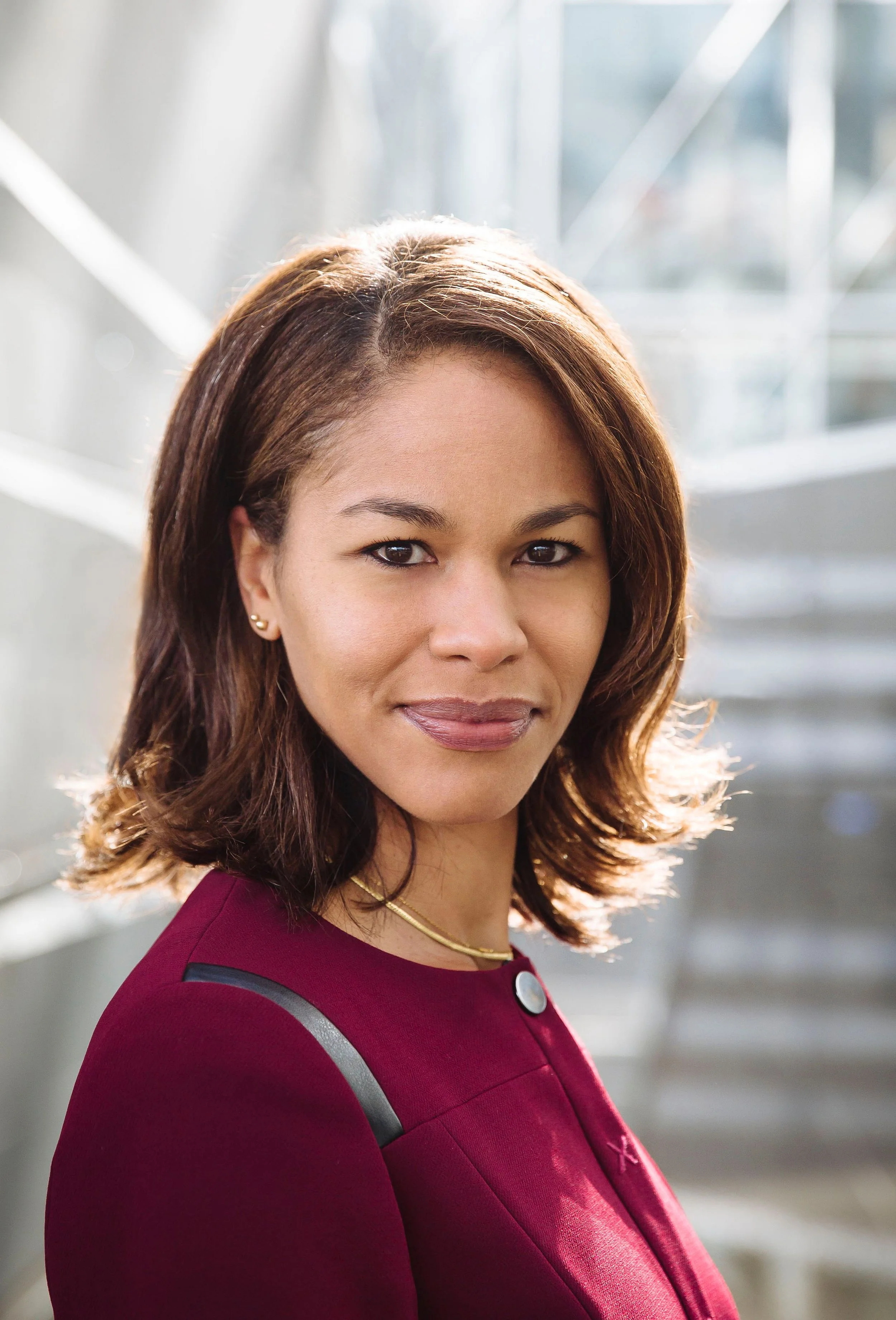 A woman with shoulder-length brown hair, wearing a burgundy blazer with black accents and a gold necklace, standing in a modern, well-lit indoor space with stairs in the background.