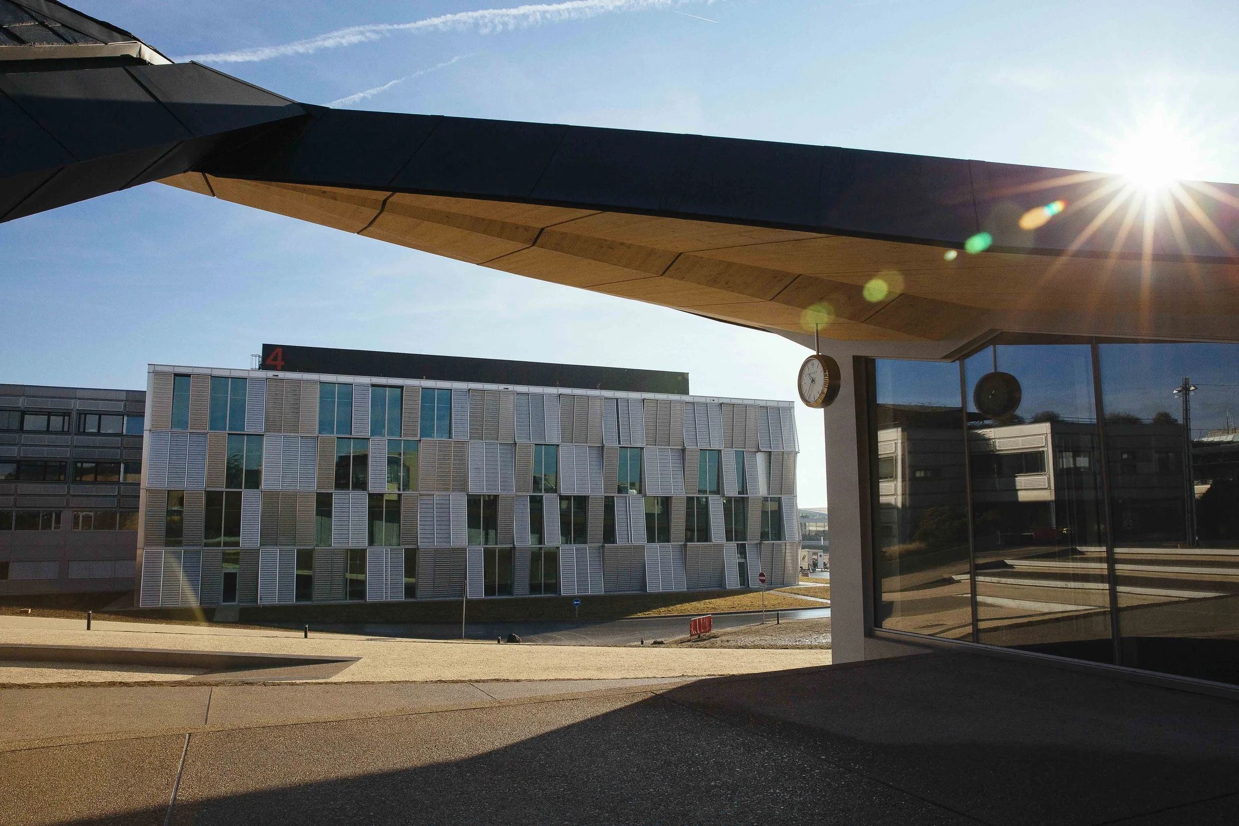 Modern building with geometric facade, seen from under a covered area with a clock and hanging lights, sunlight in the background.