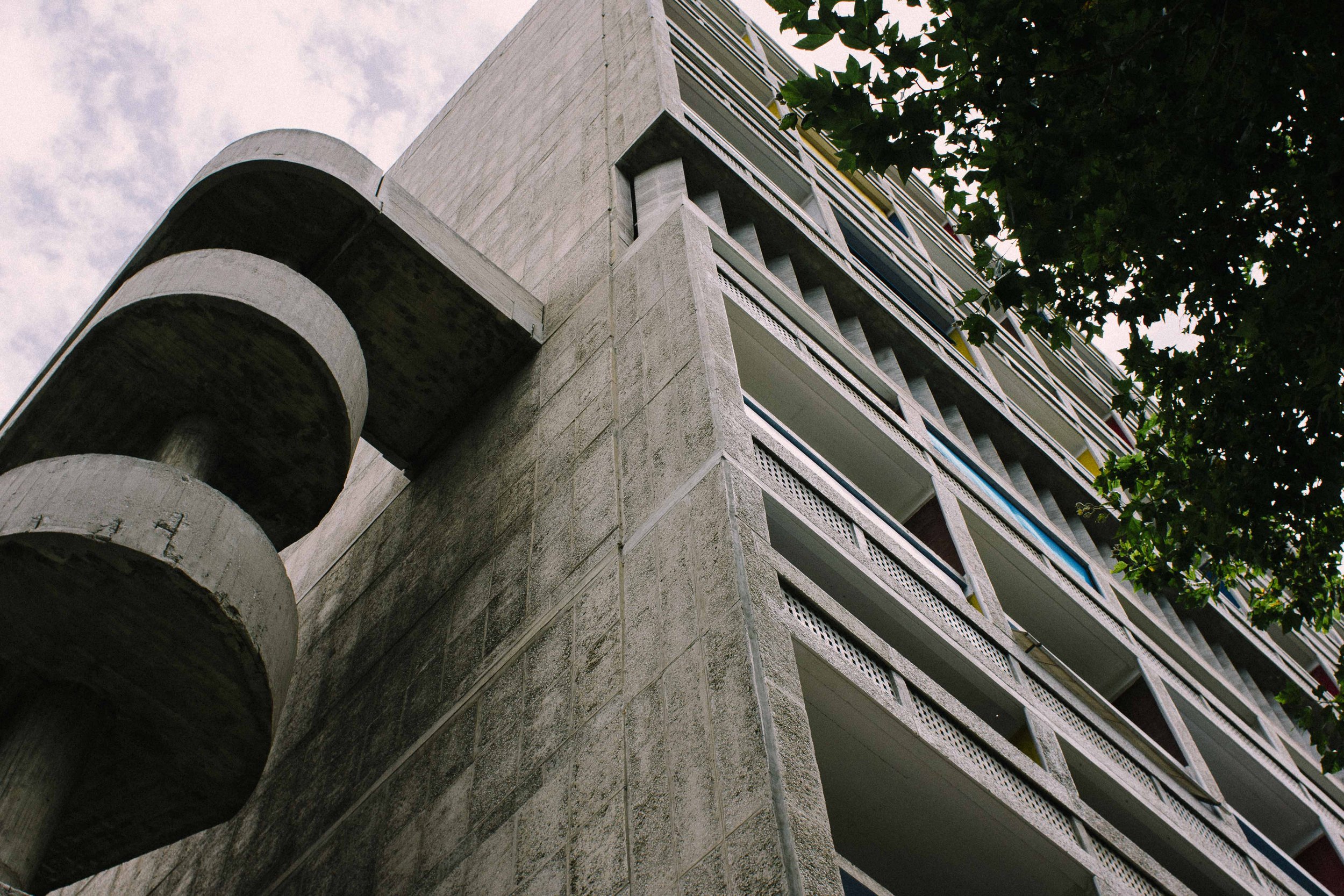 Low-angle view of a concrete building with balcony railings, partially obscured by green tree leaves in the top right corner, under a partly cloudy sky.