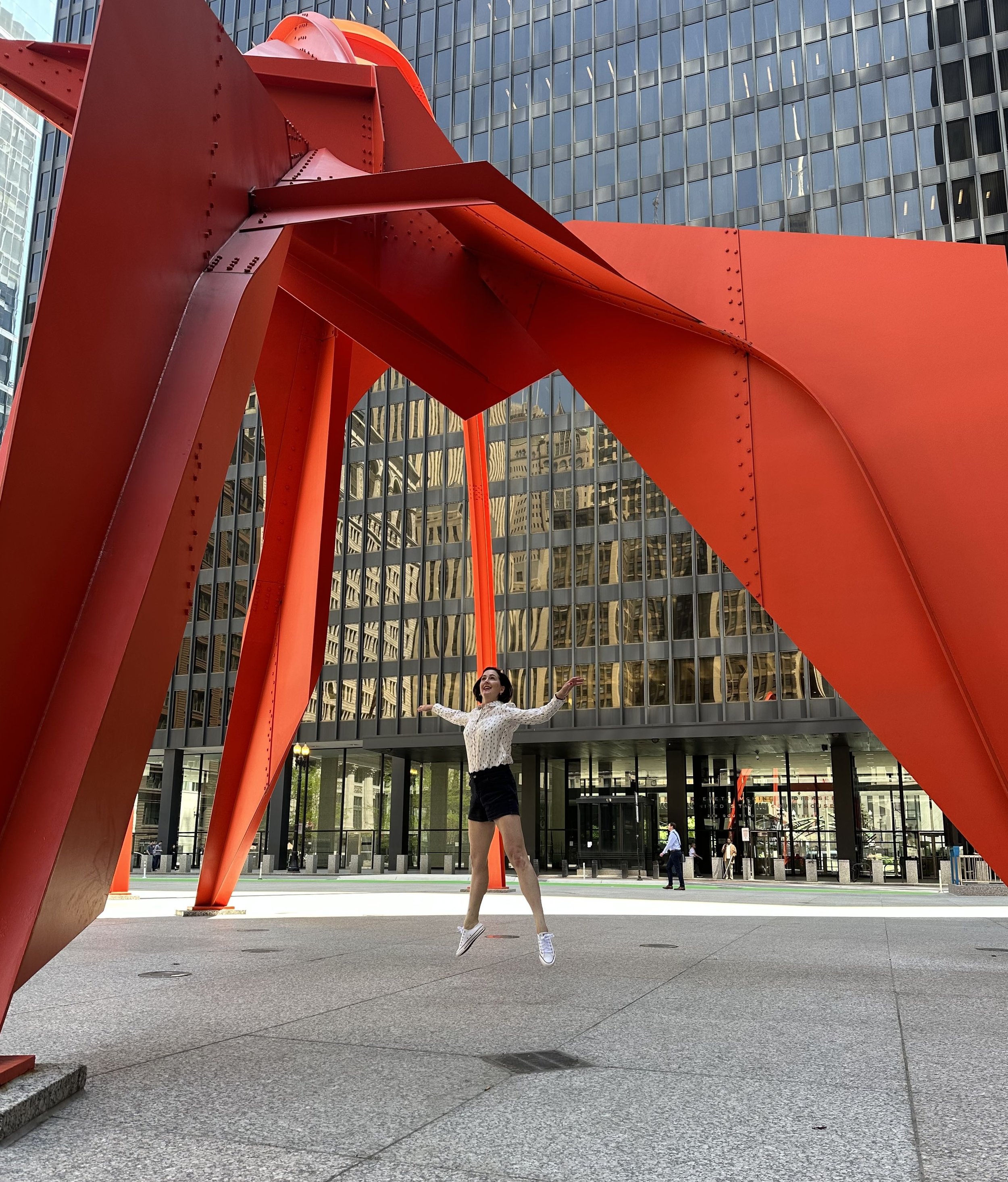 A woman jumping in front of a large abstract red sculpture in an urban setting with tall glass buildings in the background.