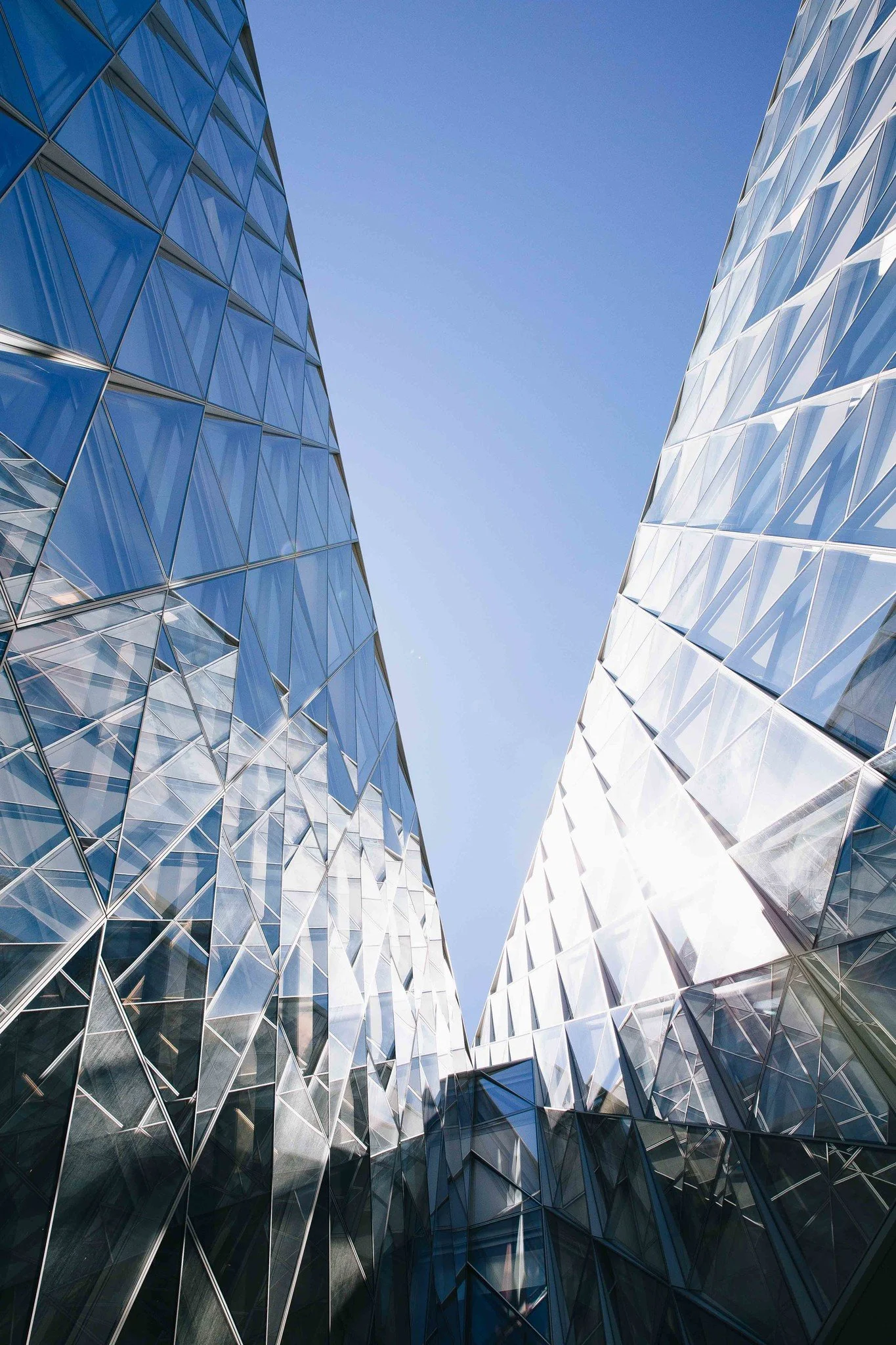 Looking up at modern glass skyscrapers with geometric facades against a blue sky.