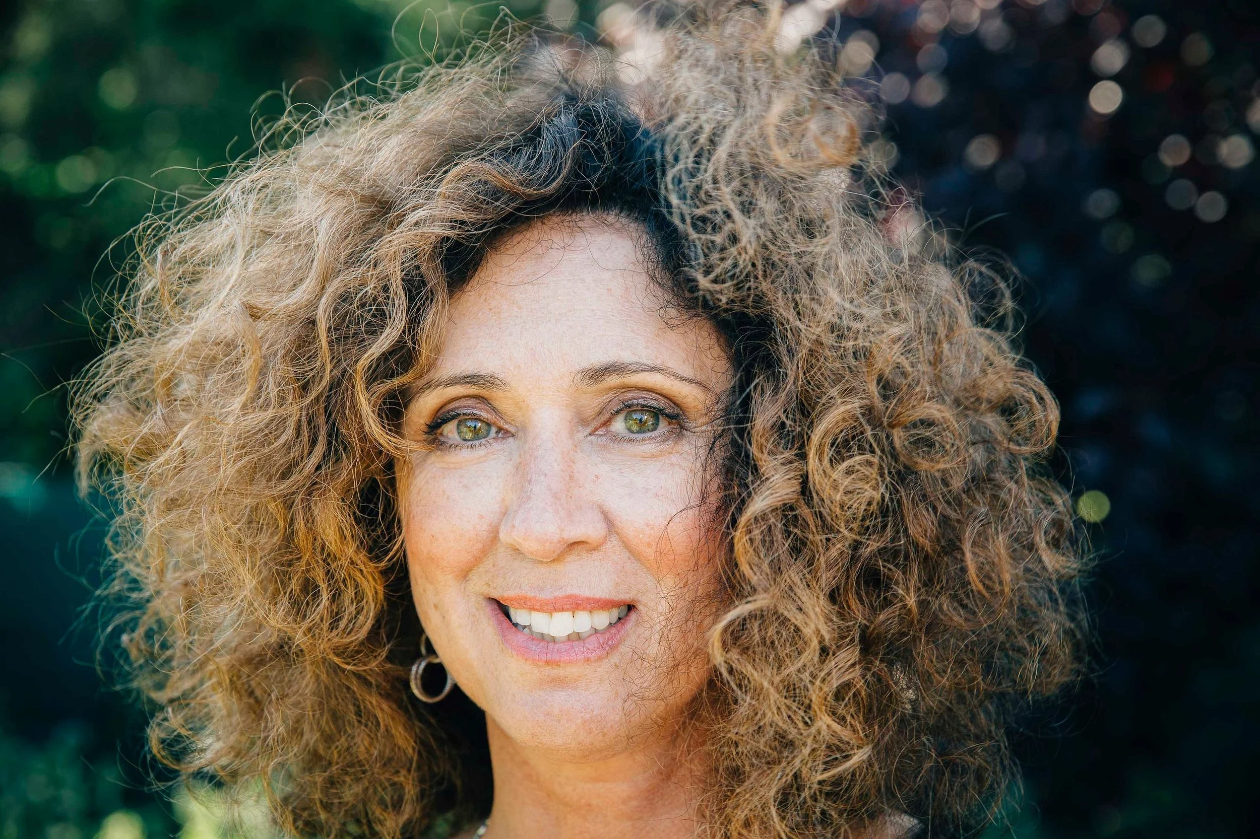 A woman with curly, voluminous hair and light-colored eyes, smiling outdoors with sunlight on her face.