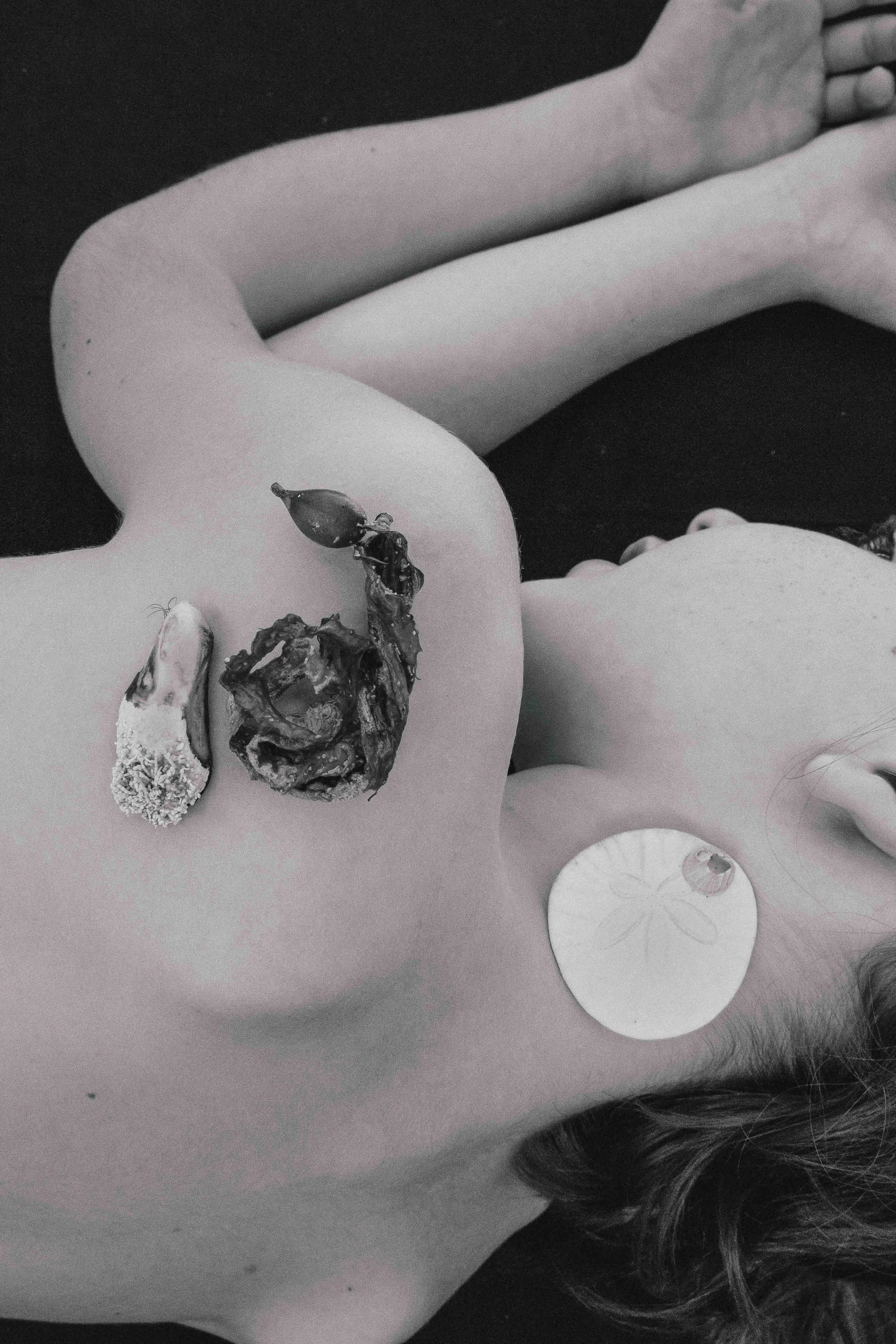 A black and white photo of a person lying down with various pieces of seashells and seaweed placed on her back and neck.