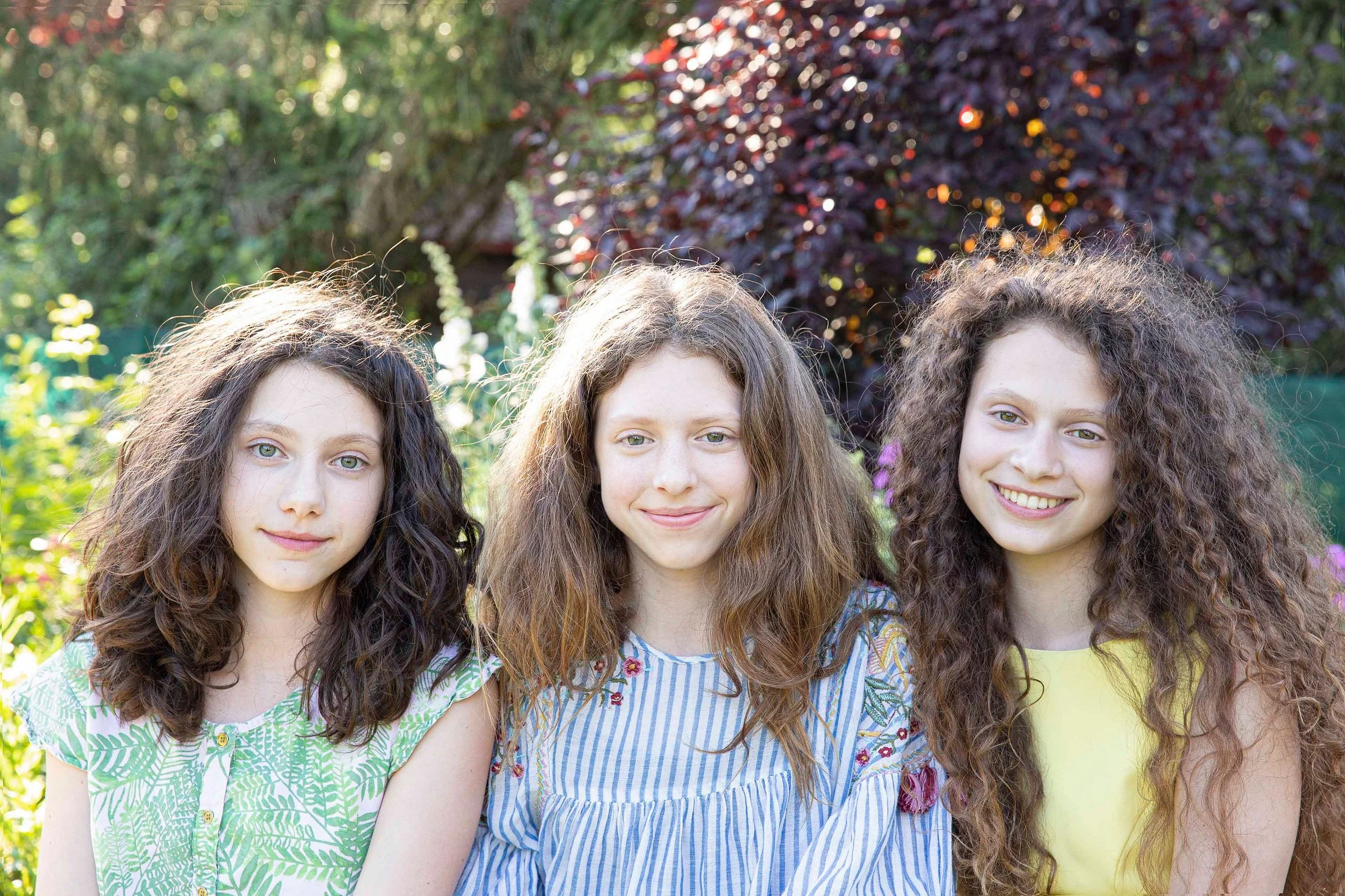 Three young girls with curly hair smiling outdoors in a garden with foliage and purple flowers in the background.