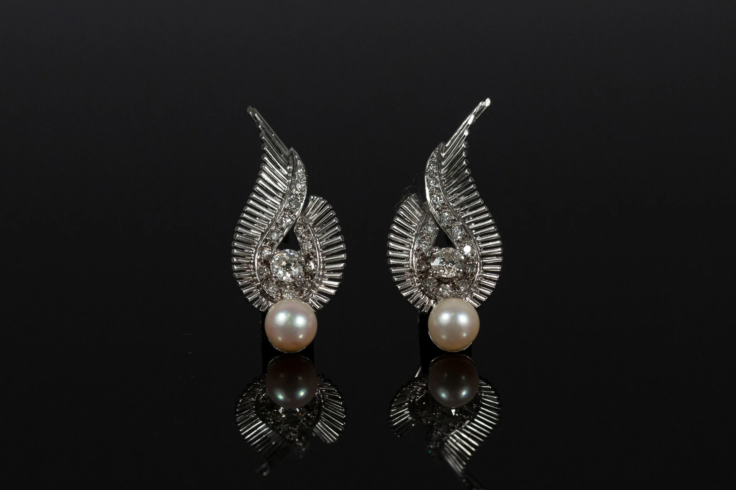 Pair of white gold earrings in art deco design with a teardrop design, encrusted with small diamonds, featuring a central round pearl on a black reflective surface.