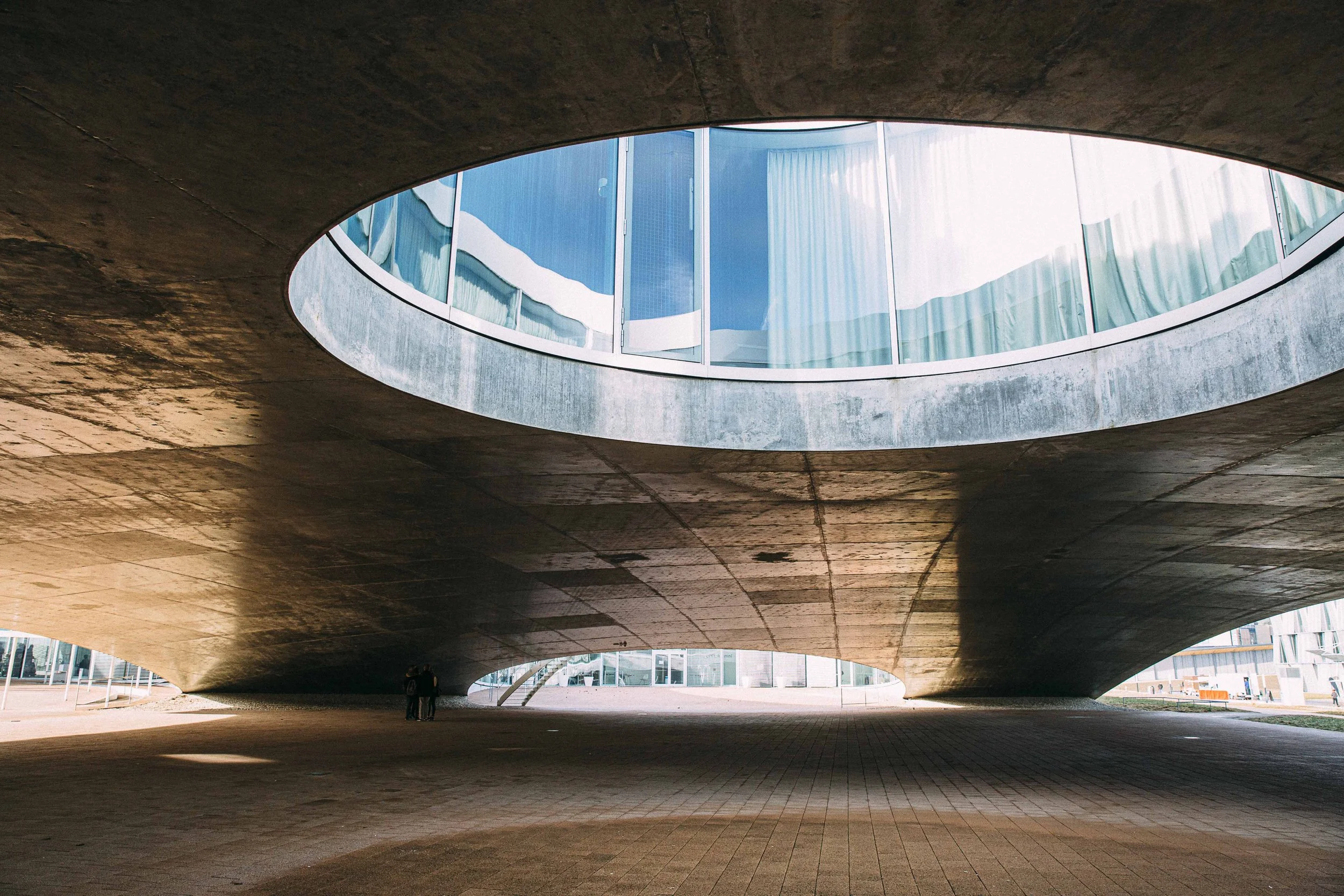 Modern structure with a large circular opening in the roof, modern building with glass windows reflected in the opening, and shadows underneath.