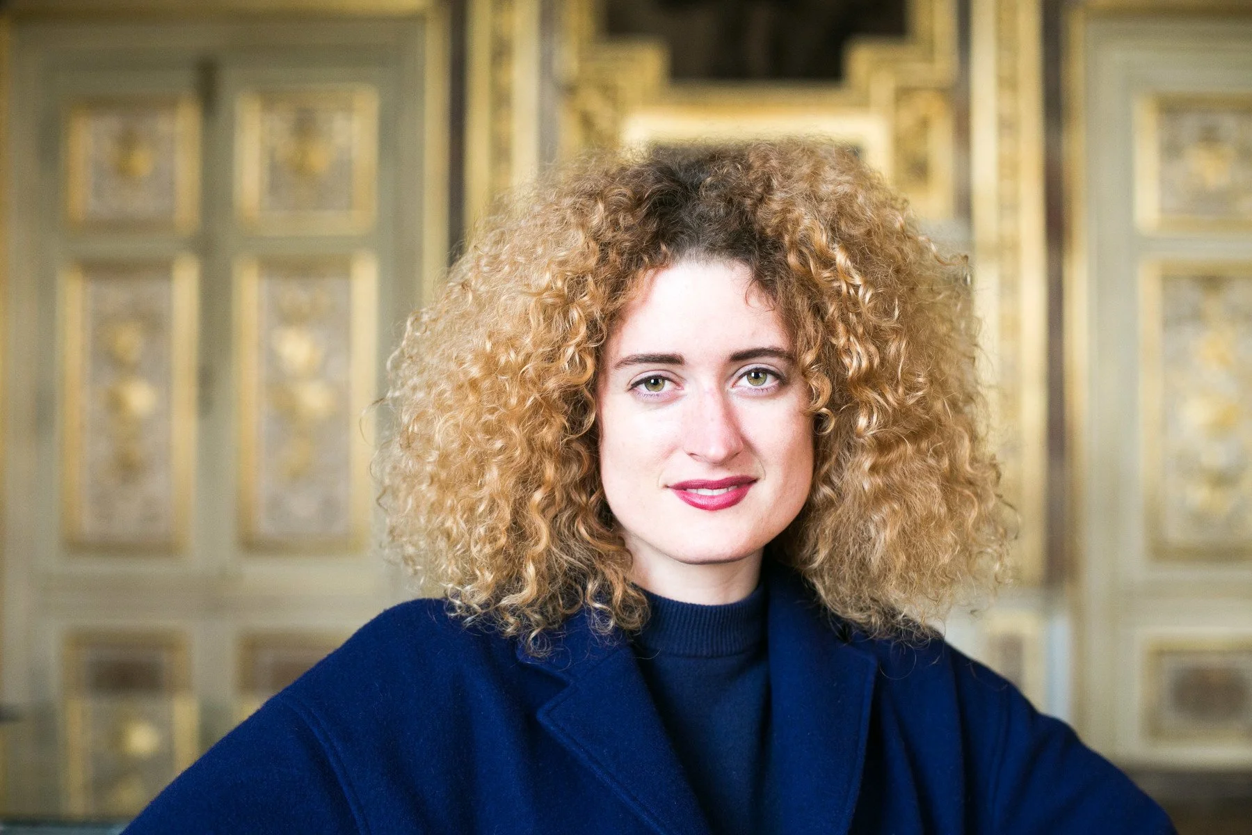 A woman with curly blonde hair wearing a dark blue blazer in the Louvre in Paris, standing in an ornate room with gold accents and intricate decor.