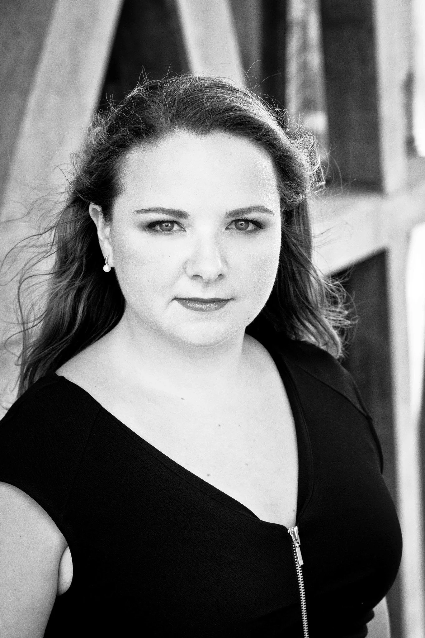 Black and white portrait of a woman with shoulder-length hair, wearing a black top with a zipper, standing outdoors with a brick building in the background.