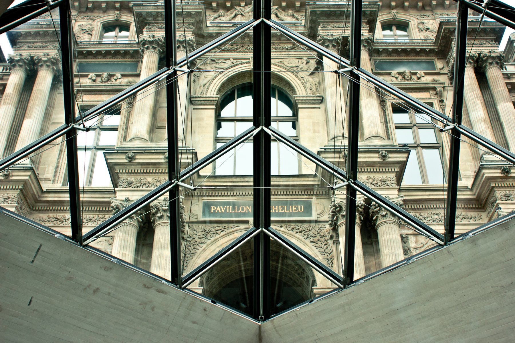 View of classical architectural building through a glass and metal structure, featuring decorative columns, sculptures, and a sign that reads 'Pavillon Chelieu'.