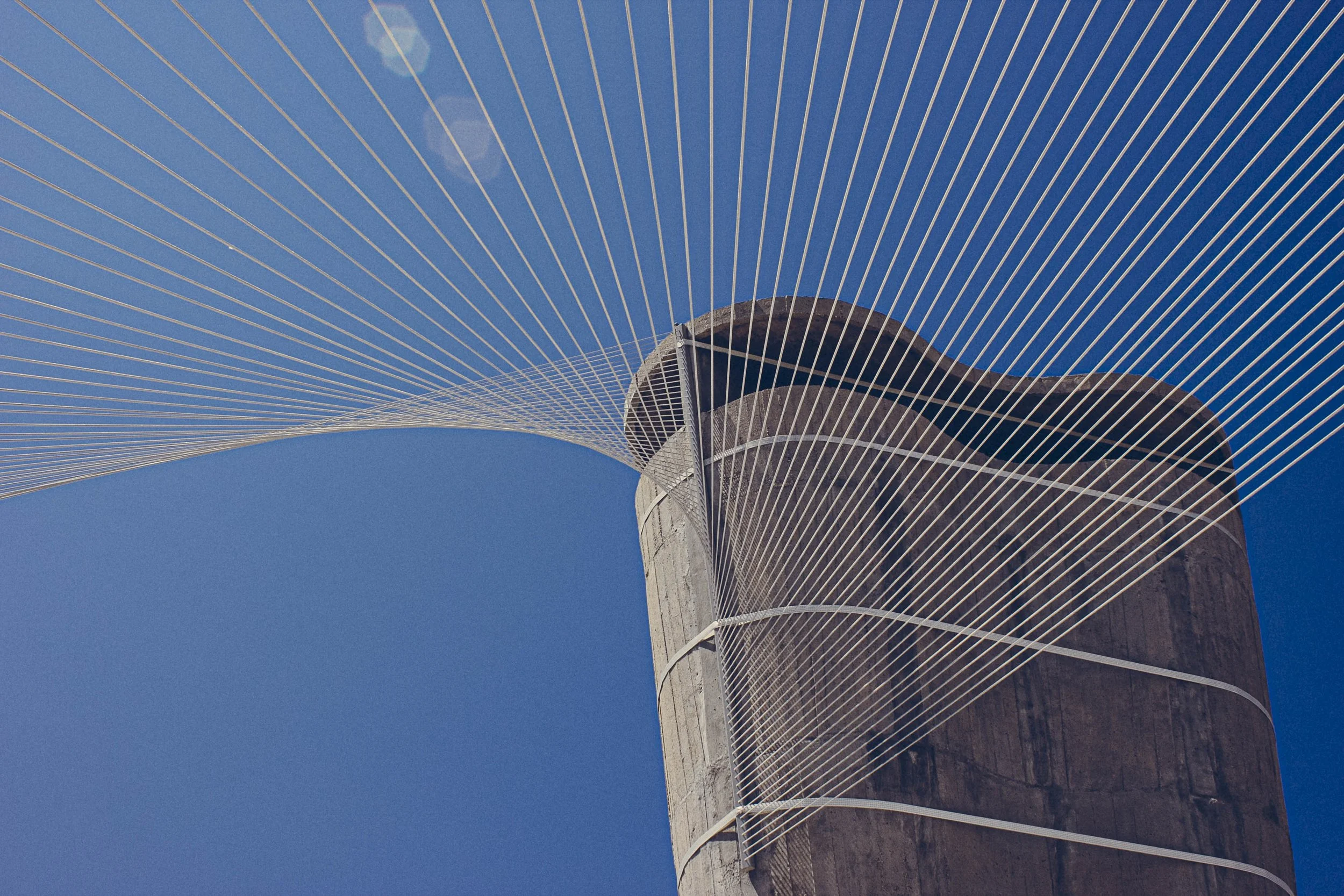 Close-up of a modern cable-stayed bridge tower with white cables radiating from the top against a clear blue sky.