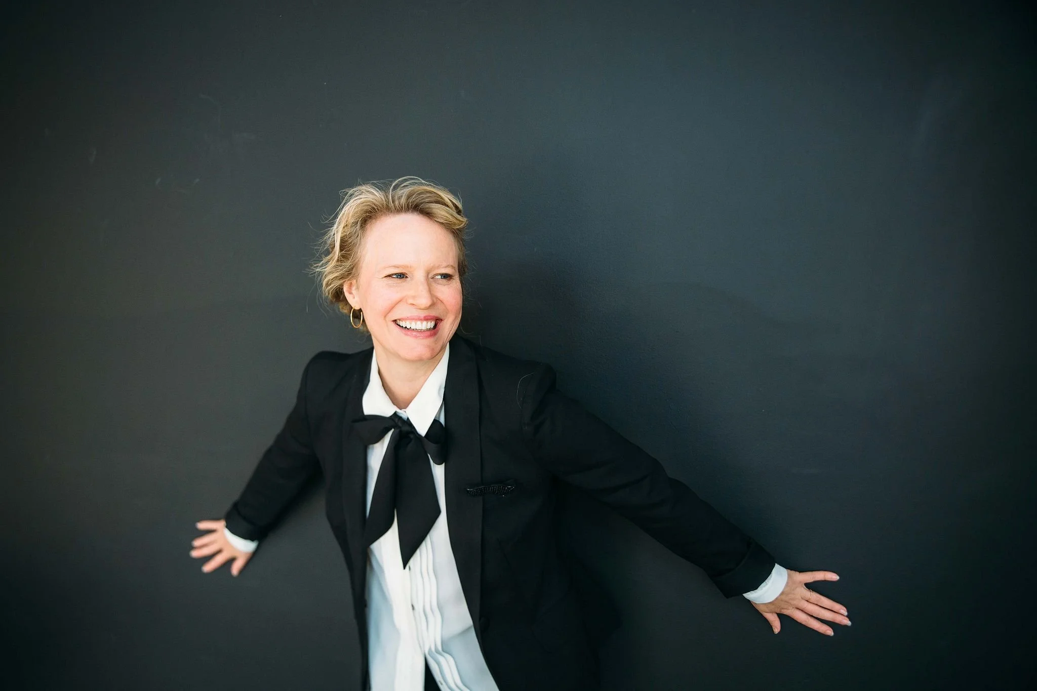 A smiling woman with blonde curly hair tied back, wearing a black blazer and a white shirt with a black bow, standing against a dark background.