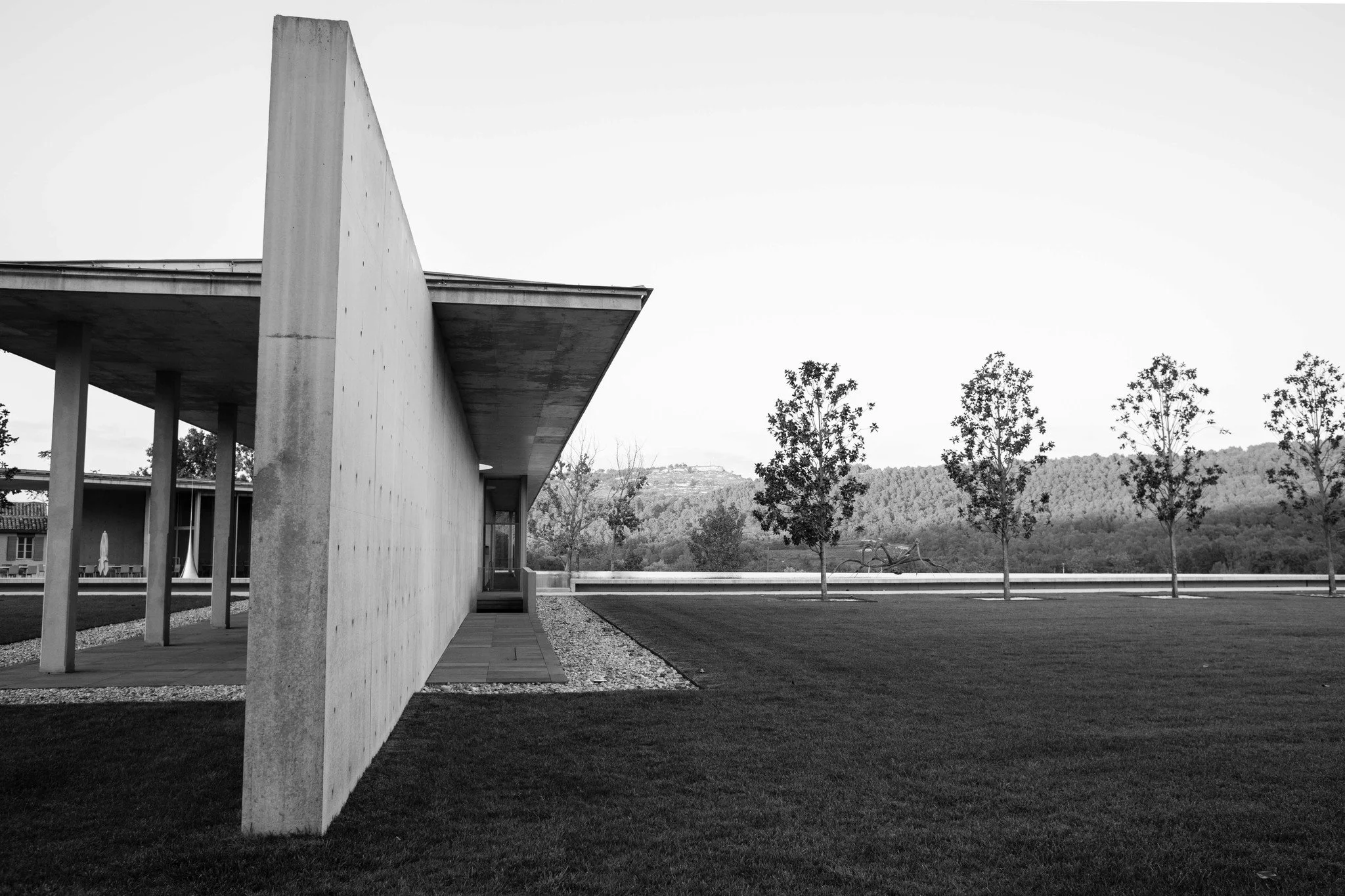 A modern building with a concrete wall and a sidewalk running alongside it, with five evenly spaced trees in the background, on a grassy field, and hills in the distance.