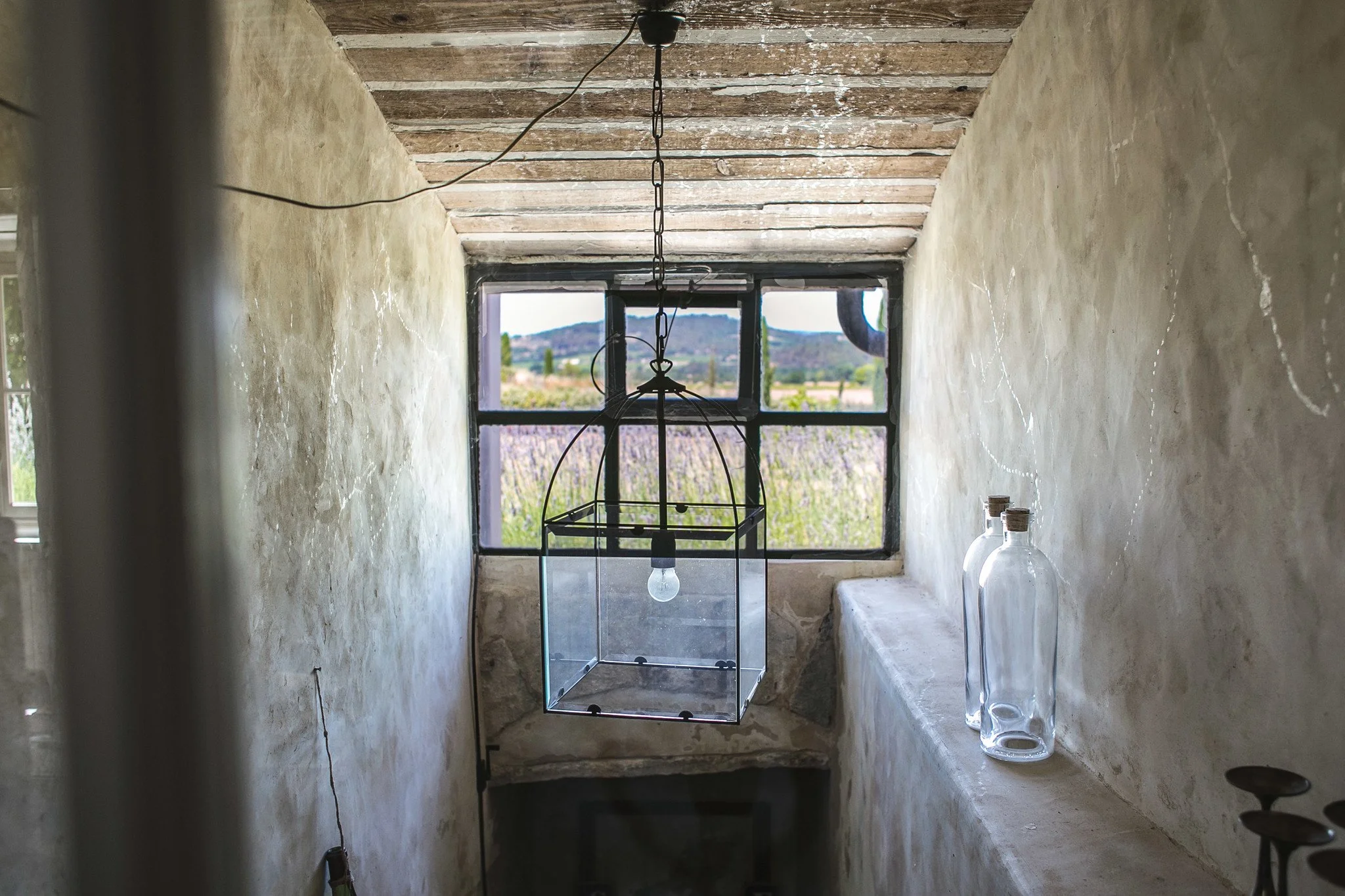 A rustic interior with plastered walls, wooden ceiling beams, and a window overlooking a scenic landscape with mountains and fields. A hanging modern lantern with a glass enclosure and a light bulb inside is suspended from the ceiling. On a ledge to 