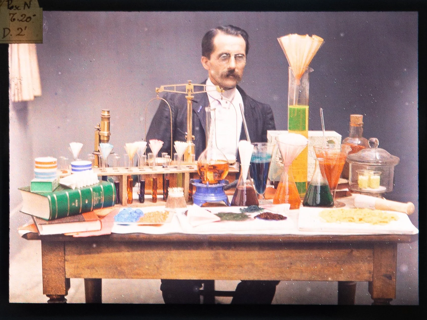 Photo of an autochrome plate with a scientist with glasses and a mustache standing behind a wooden table filled with various scientific glassware, books, and colorful chemicals, in a laboratory setting.