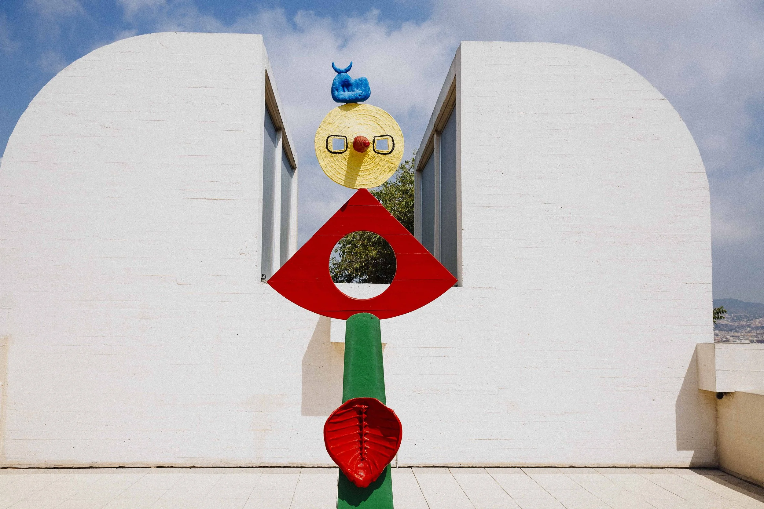 Colorful abstract sculpture with a yellow face, blue headpiece, red diamond-shaped body, and green base, placed in front of a white curved wall with a blue sky and clouds overhead.