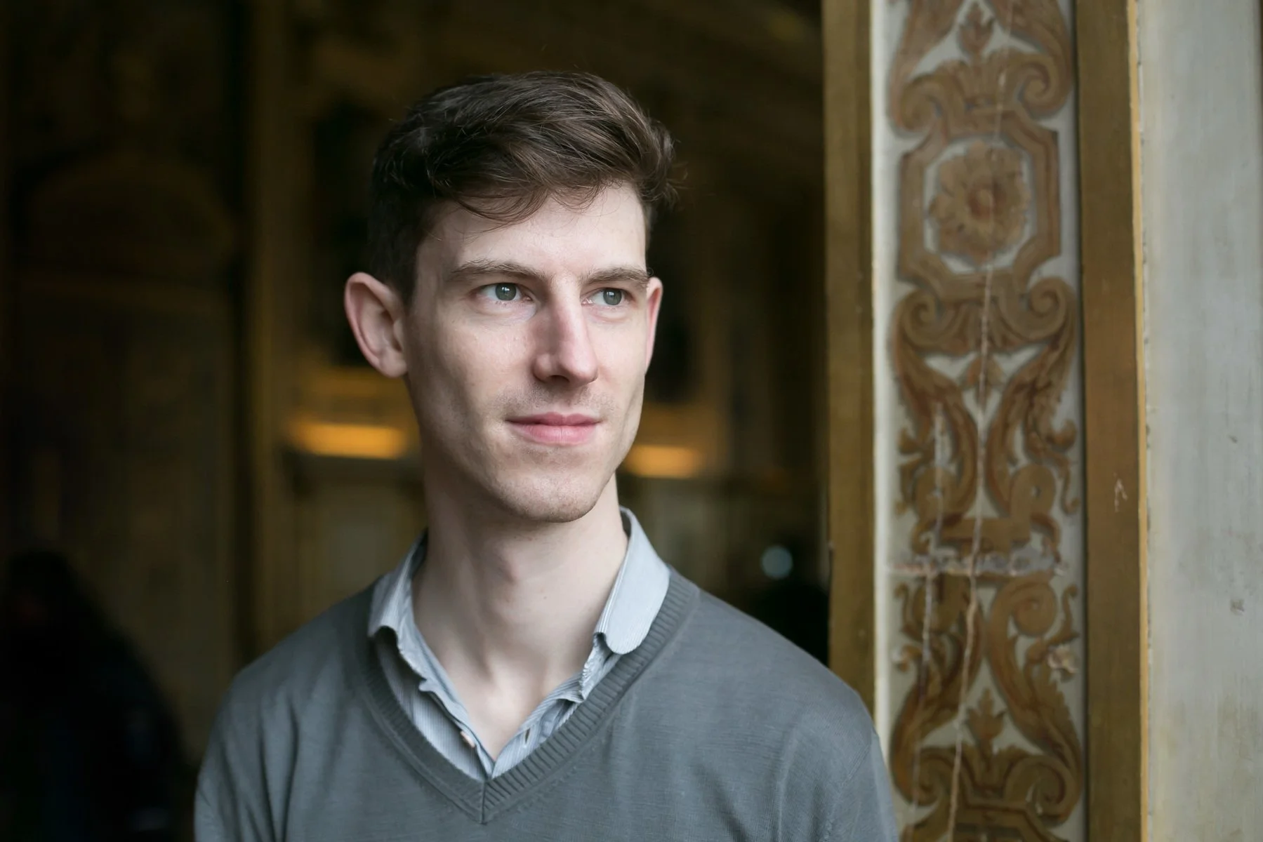A young man with brown hair and blue eyes, wearing a gray sweater over a collared shirt, standing indoors near an ornately decorated wall.