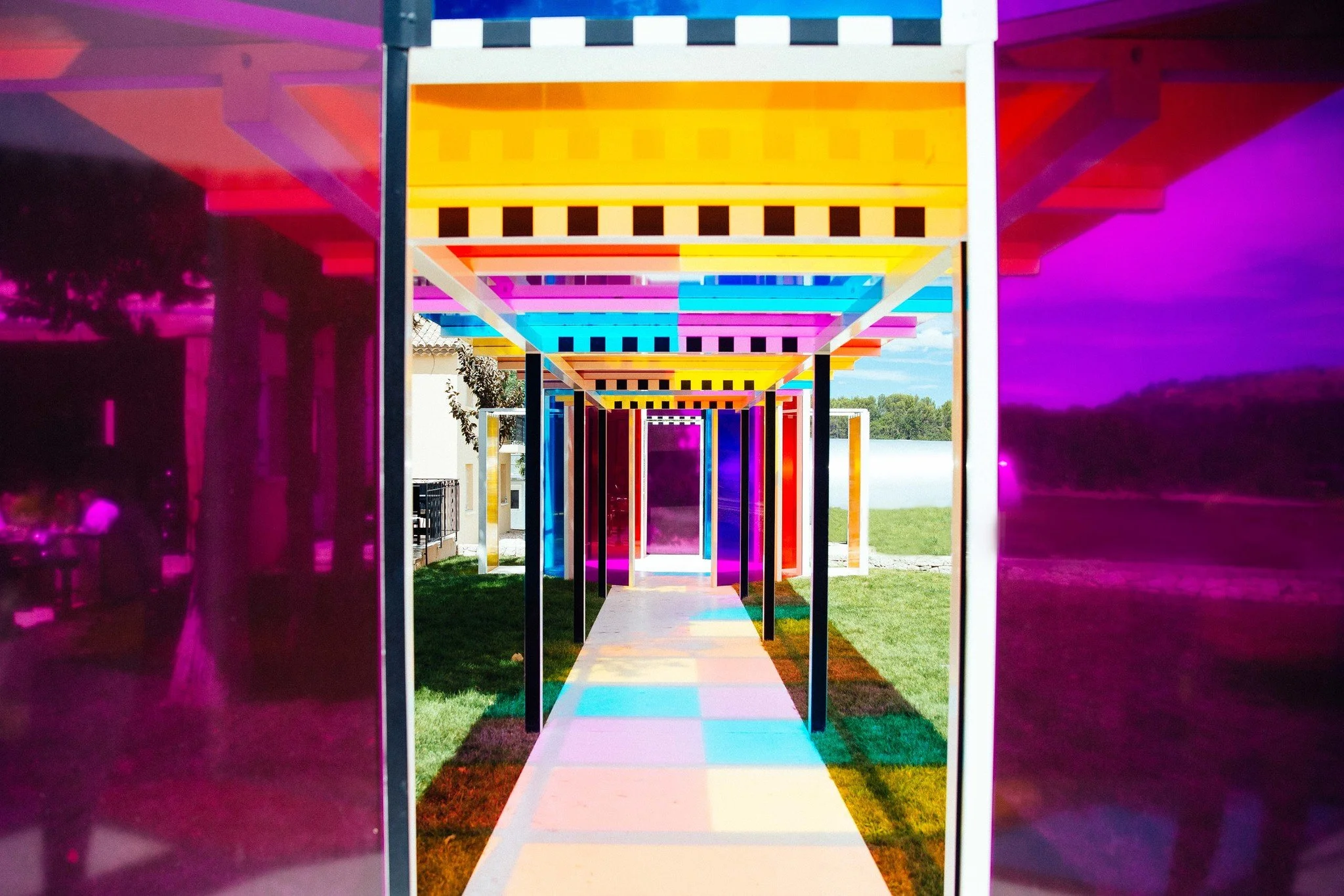 Colorful art installation featuring a corridor of geometric shapes with vibrant rainbow colors, outside in a grassy area under a partly cloudy sky.
