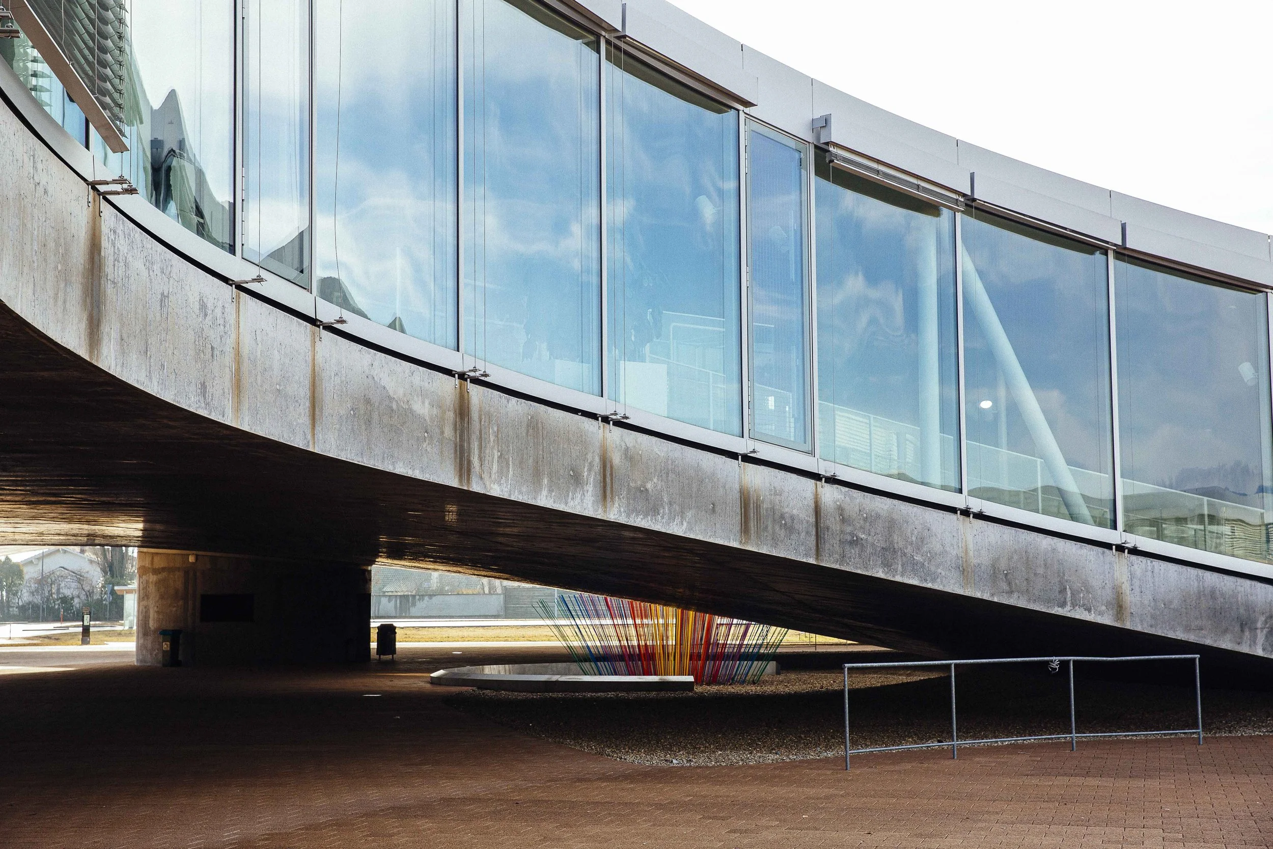 An angled view of a modern building with glass windows, a concrete underside, and a colorful sculpture underneath.