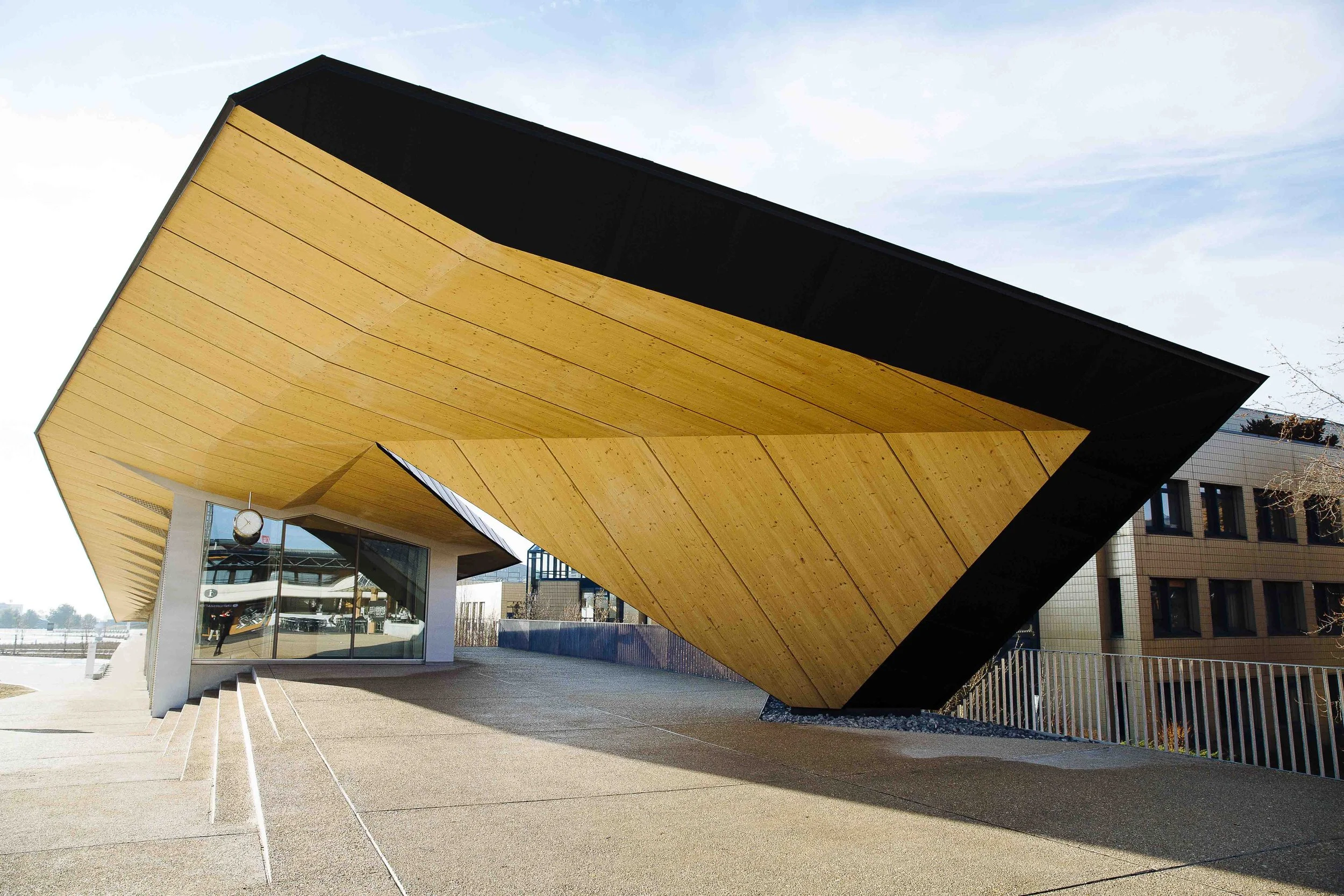 Modern building with an overhanging wooden roof, glass entrance, and a clock outside.