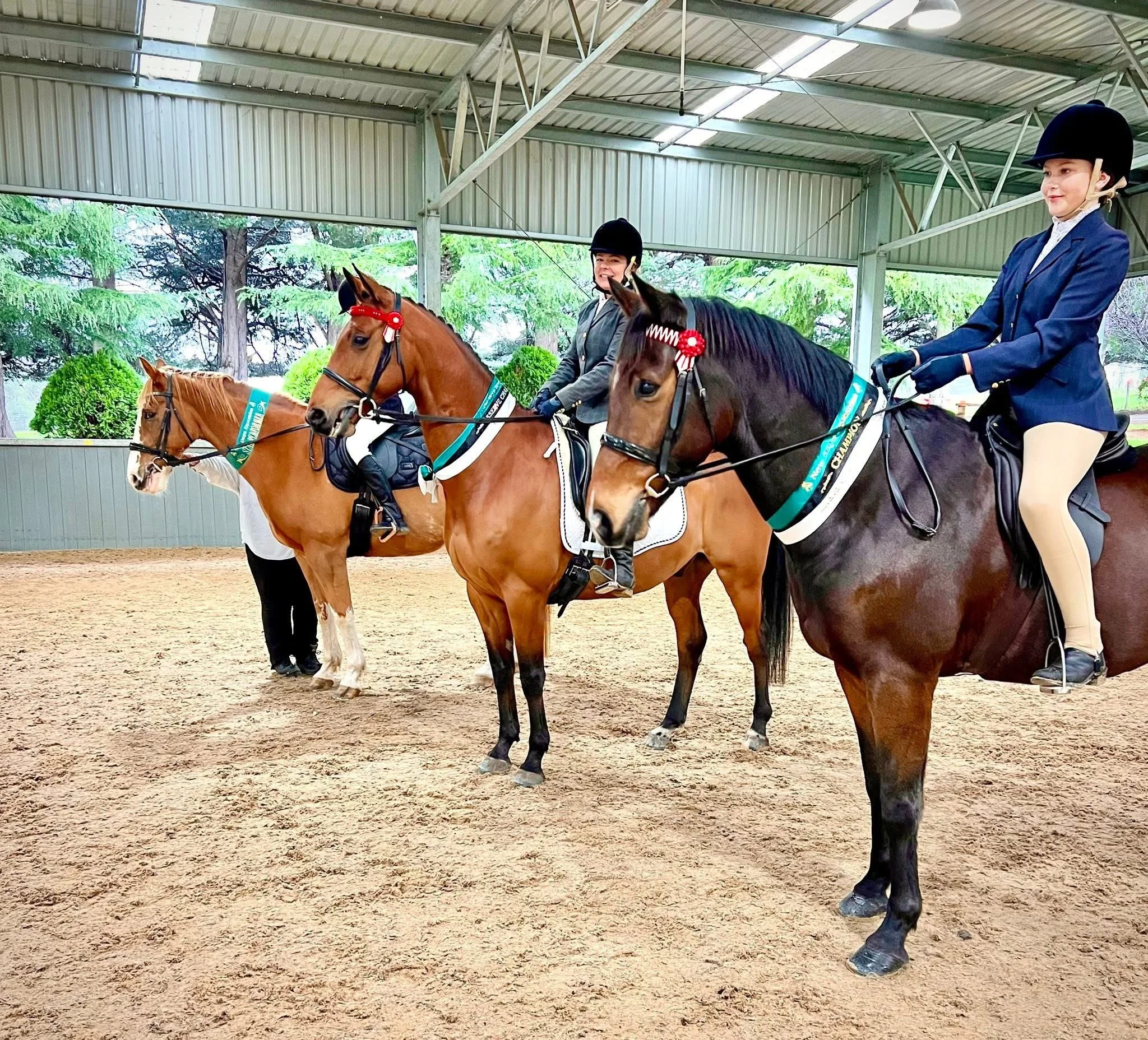 Two young women in riding attire riding horses inside an indoor riding arena. The women are wearing helmets and formal riding jackets, with the horses decorated with ribbons, indicating a competition or event.