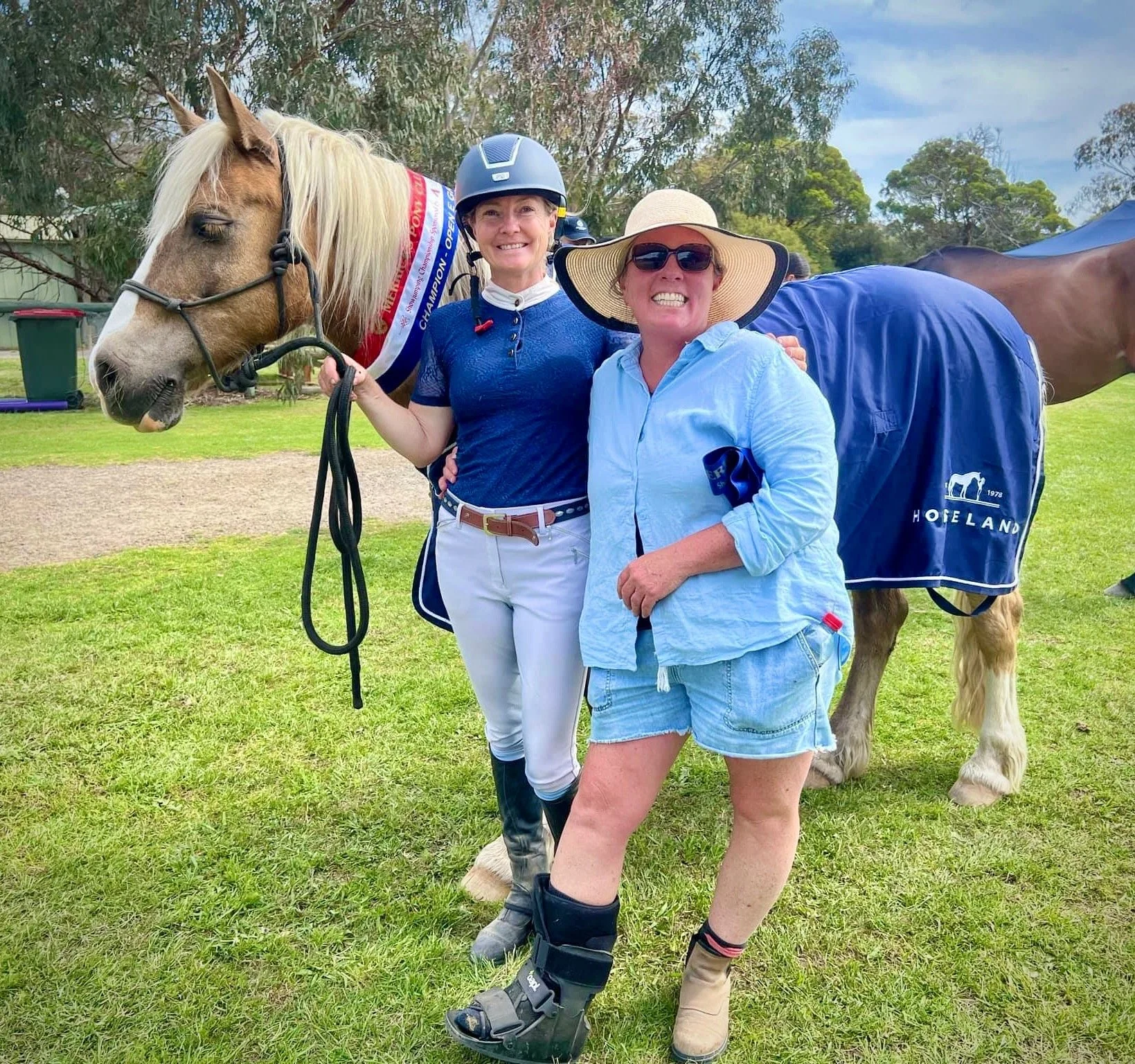 Two women smiling and standing in a field with a horse. One woman is in riding gear and helmet, holding the horse's bridle. The other woman is wearing a sun hat, sunglasses, a light blue shirt, and shorts.