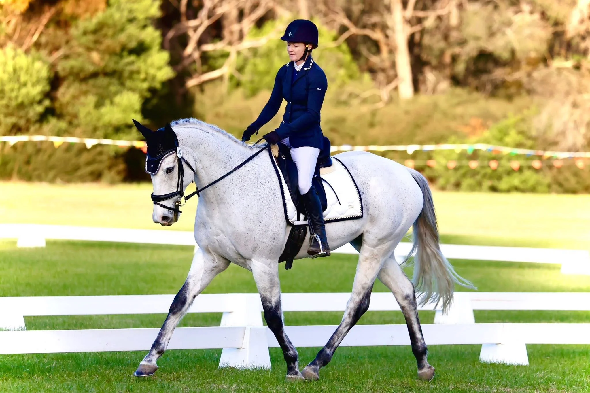 A woman in riding attire and a helmet riding a gray horse with black accents on a grassy field during a sunny day.