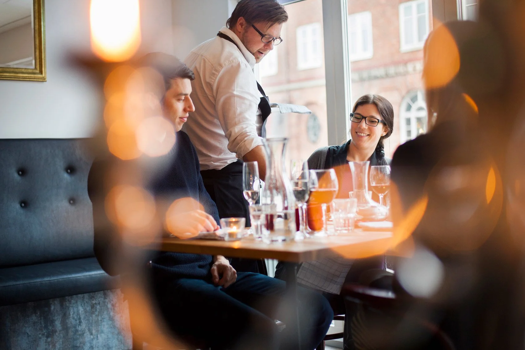 A waiter serving four customers at a restaurant table, with three women and one man, in a bright room with large windows.