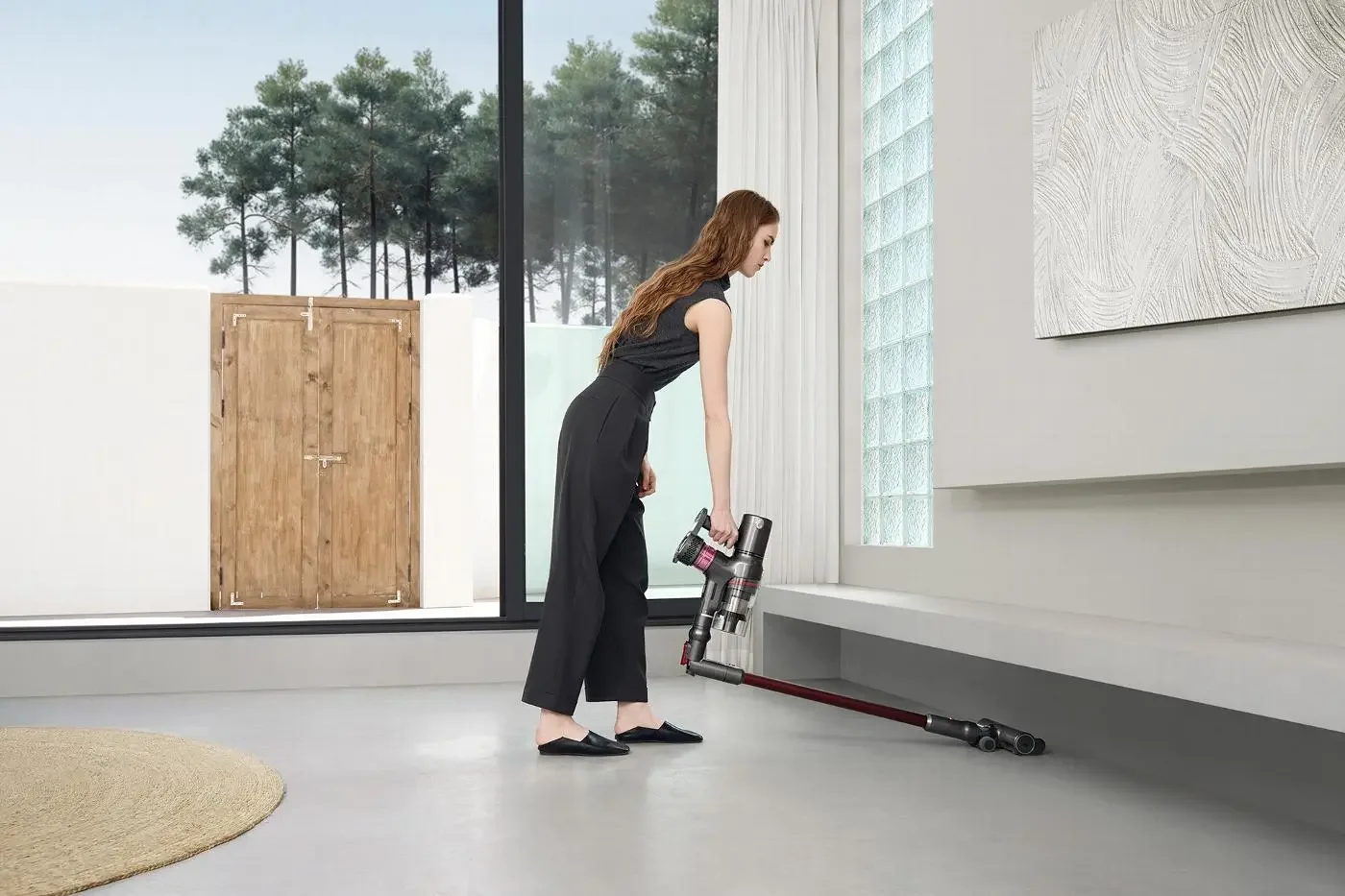 Woman vacuuming the floor inside a modern home with large window and artwork on the wall.