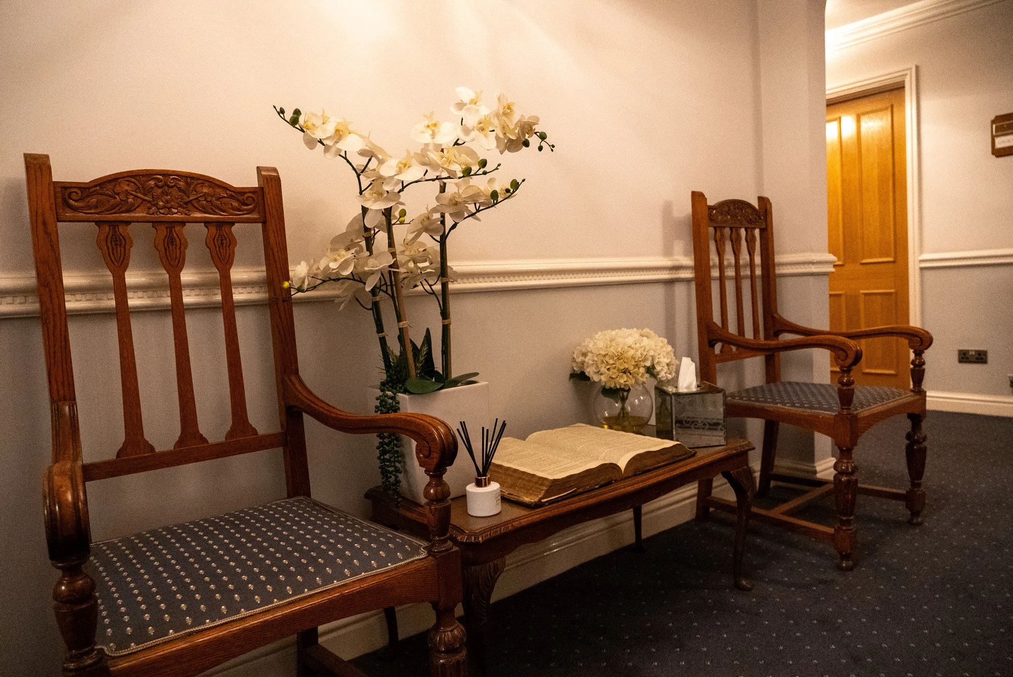 A waiting area with two wooden chairs, a floral arrangement, an open book, and a decorative table in a room with beige walls and a dark carpet.