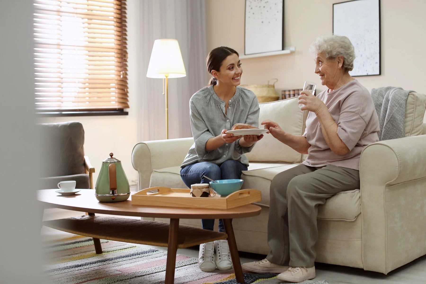 A young woman offers a plate with bread to an elderly woman sitting on a cream-colored sofa, holding a glass of water, in a cozy living room.