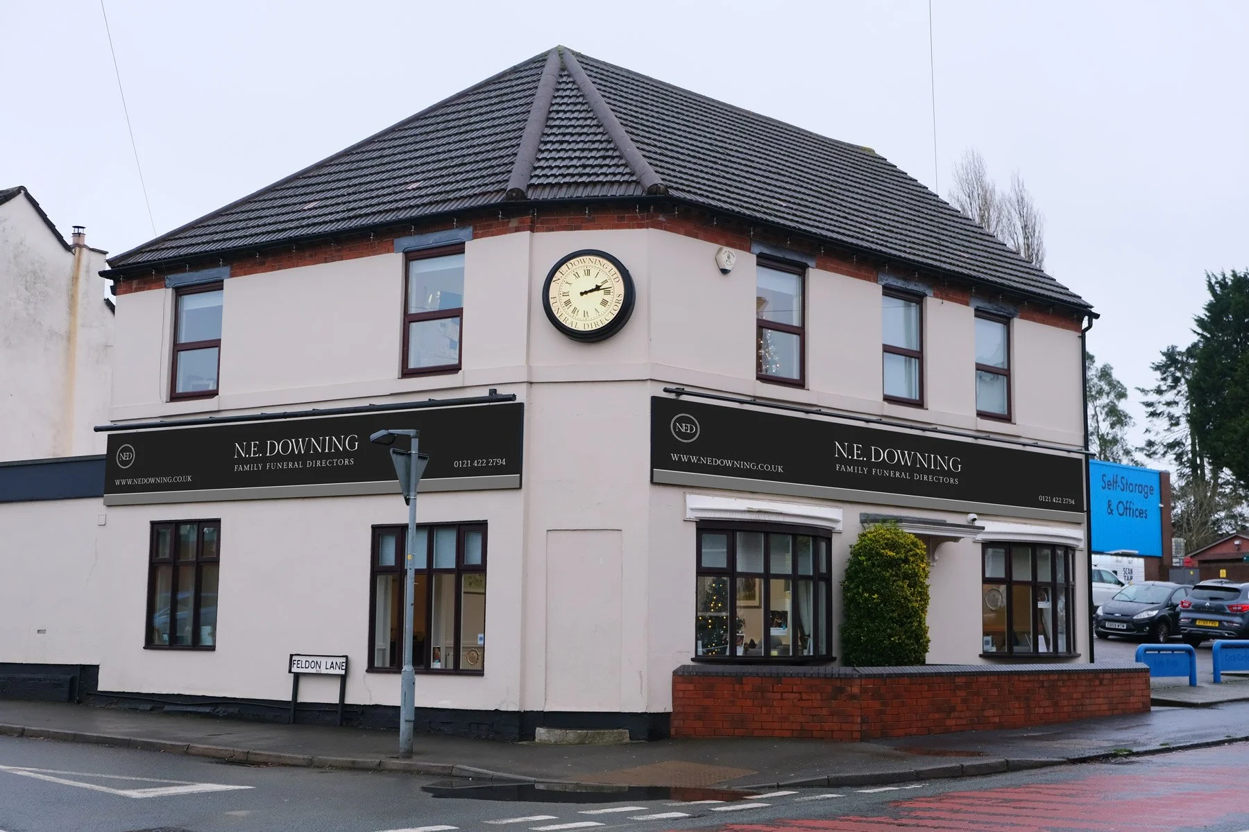 A two-story commercial building with a white facade and dark window frames, located on a street corner. The building has signage for "N.E. Downing Family Funeral Directors" and a round clock on the upper corner. There is a bush in front of the corner bump-out, a street sign reading "Feldon Lane," and parking lot with cars in the background.