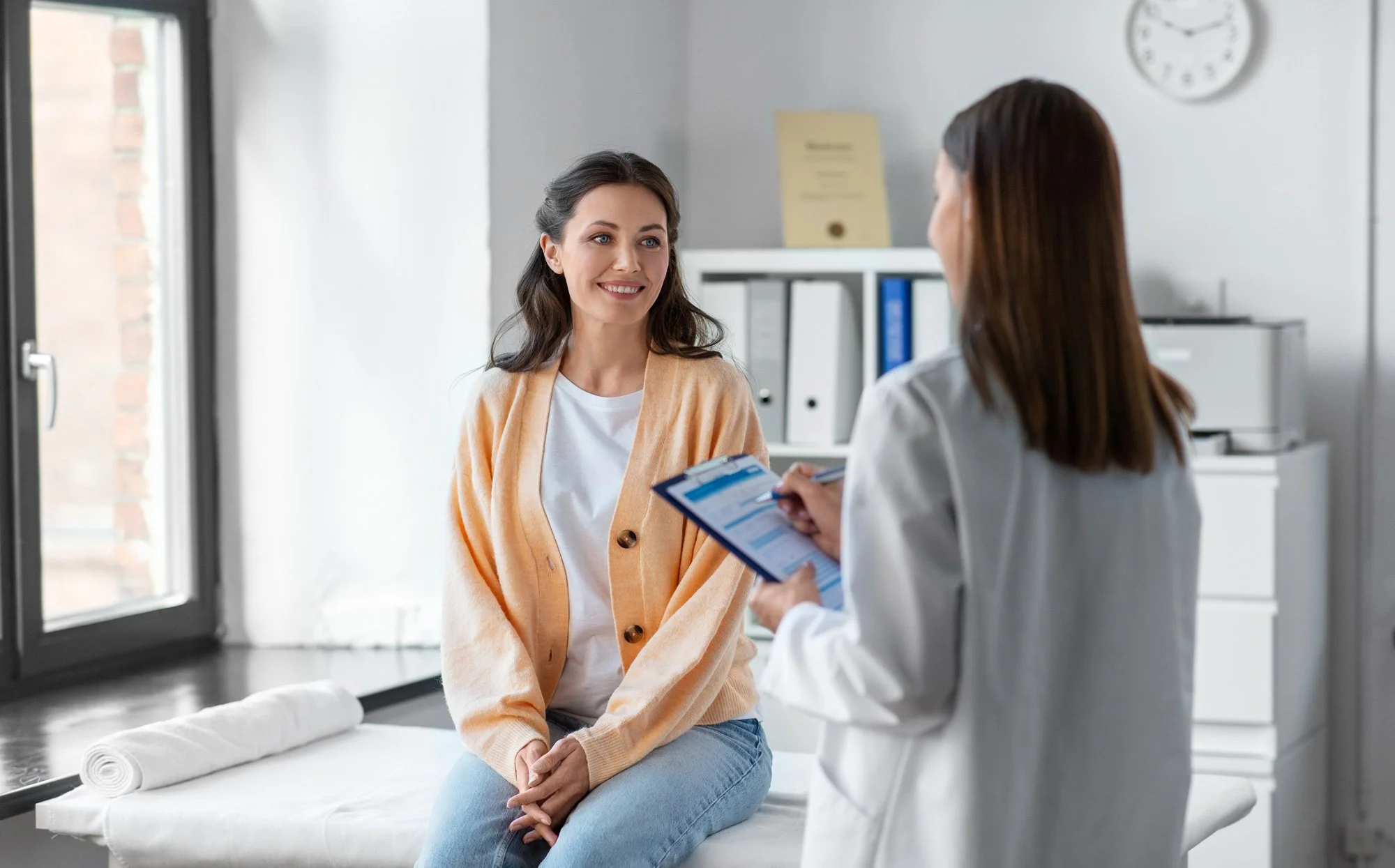 A female patient sitting on an examination table in a medical office, talking to a female healthcare provider who is holding a clipboard. The room has a window, a clock, and medical files in the background.