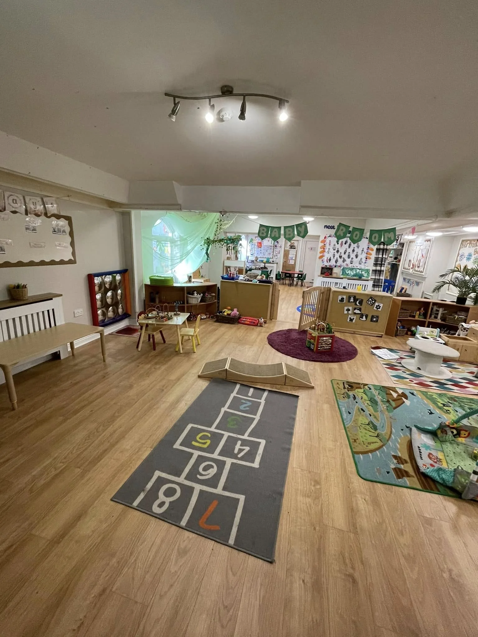 Interior view of a children's playroom with wooden flooring, a hopscotch rug, a balance beam, and various toys and play areas, decorated for a baby room.