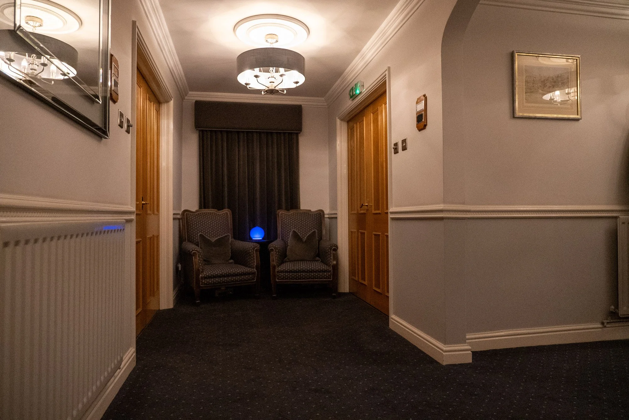 A hallway with two armchairs at the end, a small table with a blue glowing orb, framed pictures on the walls, carpeted floor, wooden doors, ceiling lights, and curtains in the background.