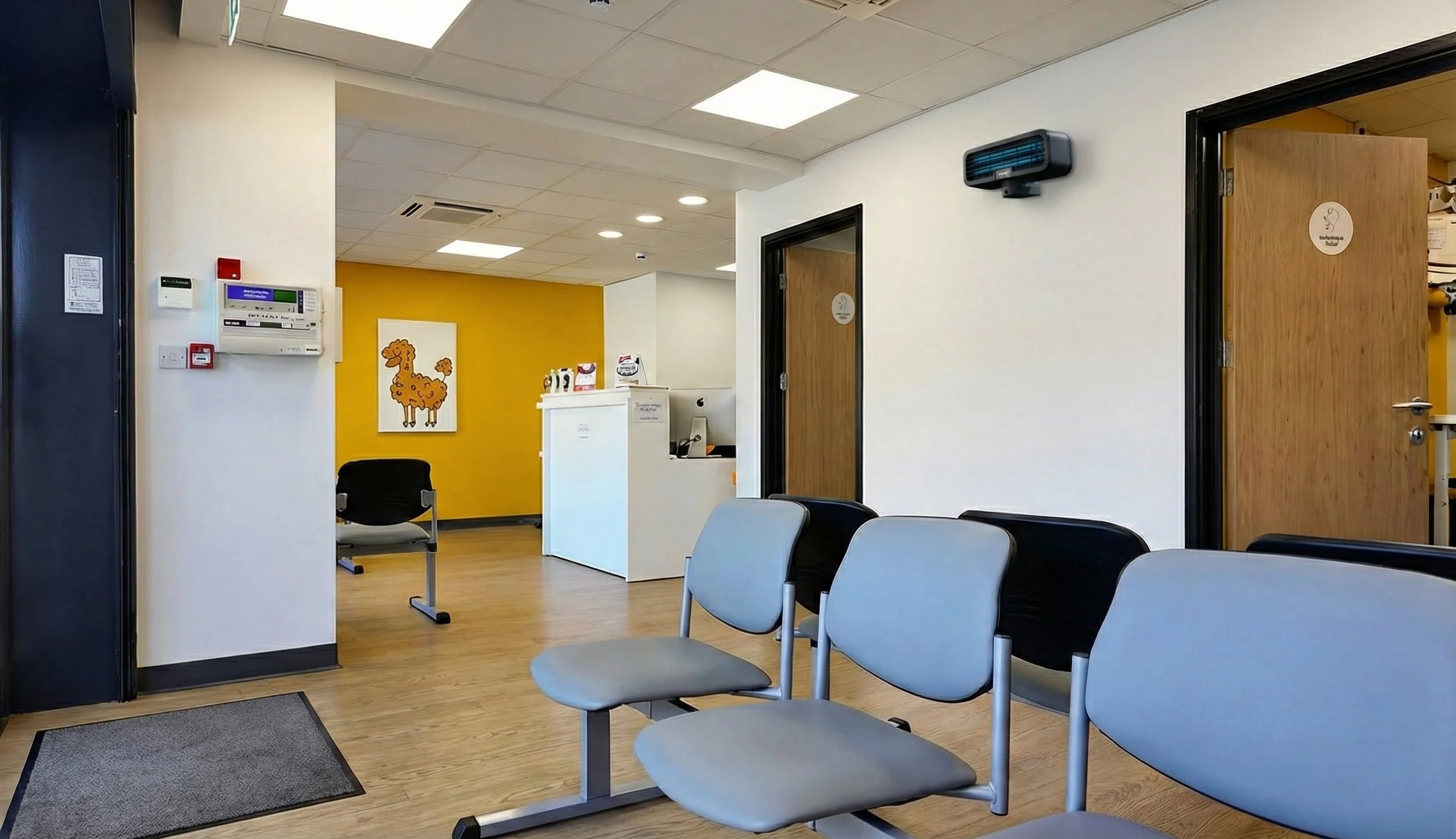 Waiting area in a medical office with four gray chairs, a dark doorway with a sign, white walls, a white reception desk, and a yellow accent wall with a giraffe painting.