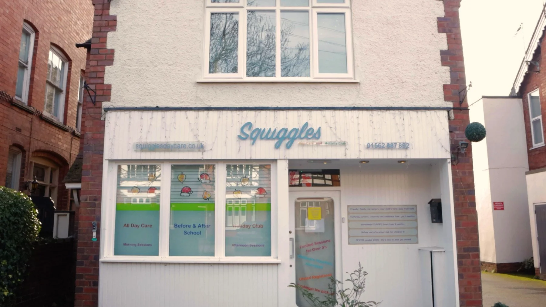 Exterior of a childcare center named Squuggles with white walls and large windows, decorated with stickers of fruits and children playing, and signs indicating services like all-day care, before and after school, holiday club, and funded sessions.