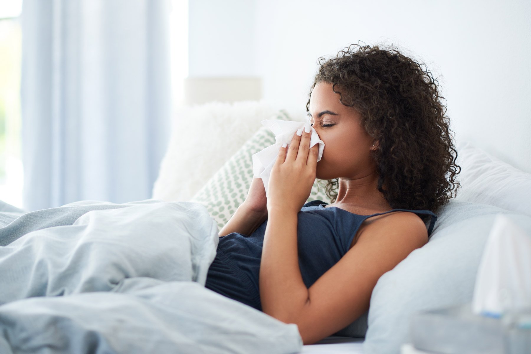 Woman lying in bed, holding a tissue to her nose, appearing to be sick or experiencing cold symptoms.