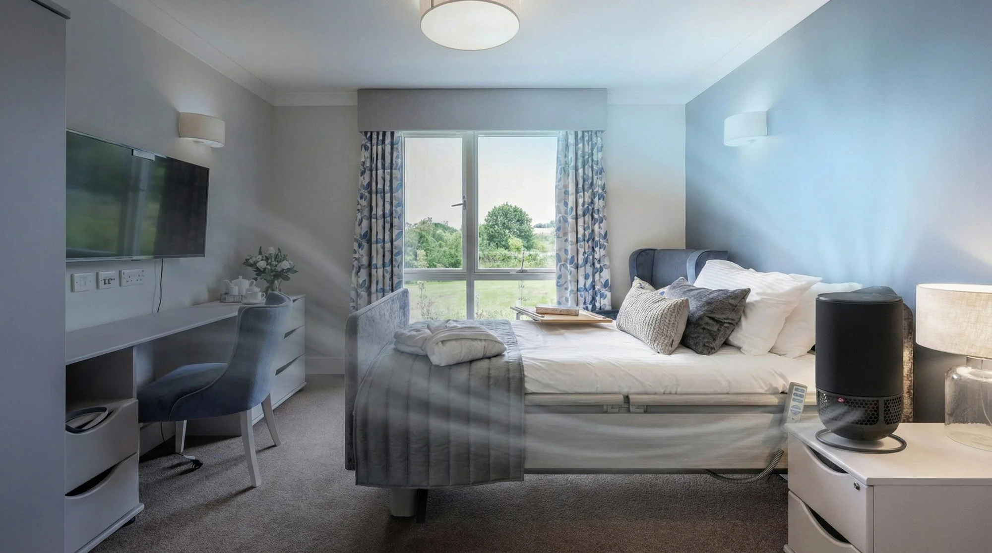 A bedroom with a bed, pillows, bedside table, TV on the wall, desk with chair, and window with curtains showing a green landscape outside.