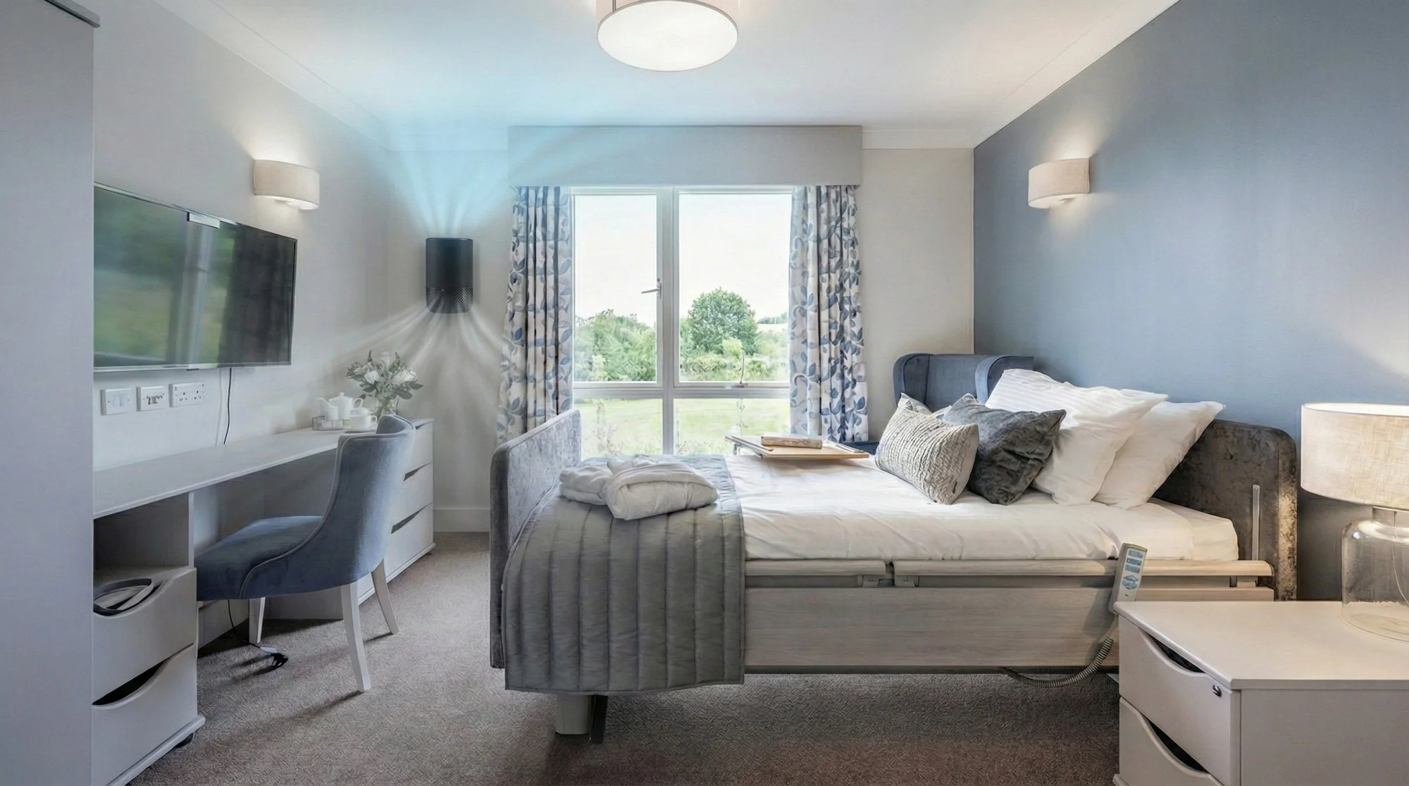 Hotel room with a bed, pillows, a desk with a chair, a wall-mounted TV, a window with blue and white patterned curtains, and a view of greenery outside.