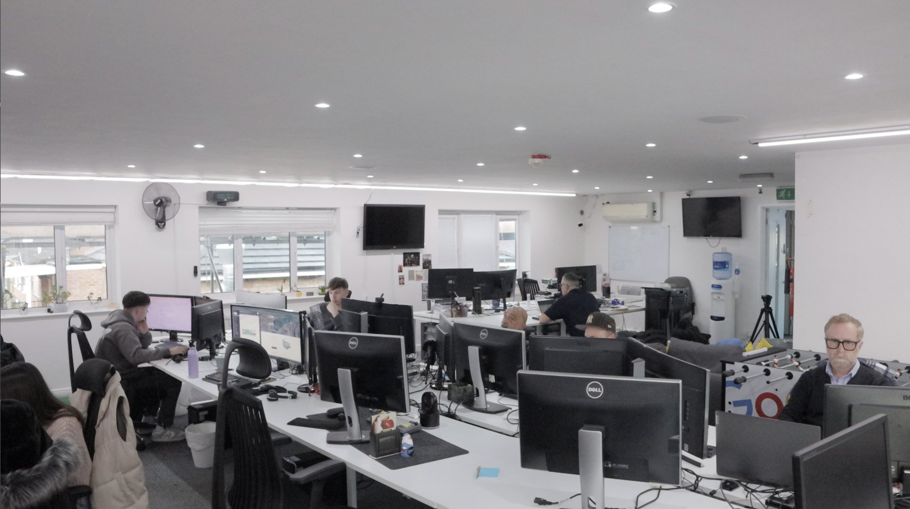 Office workspace with multiple desks, monitors, and employees working at computers. Windows on the left side, with some LED lights on the ceiling, and several TVs on the walls.