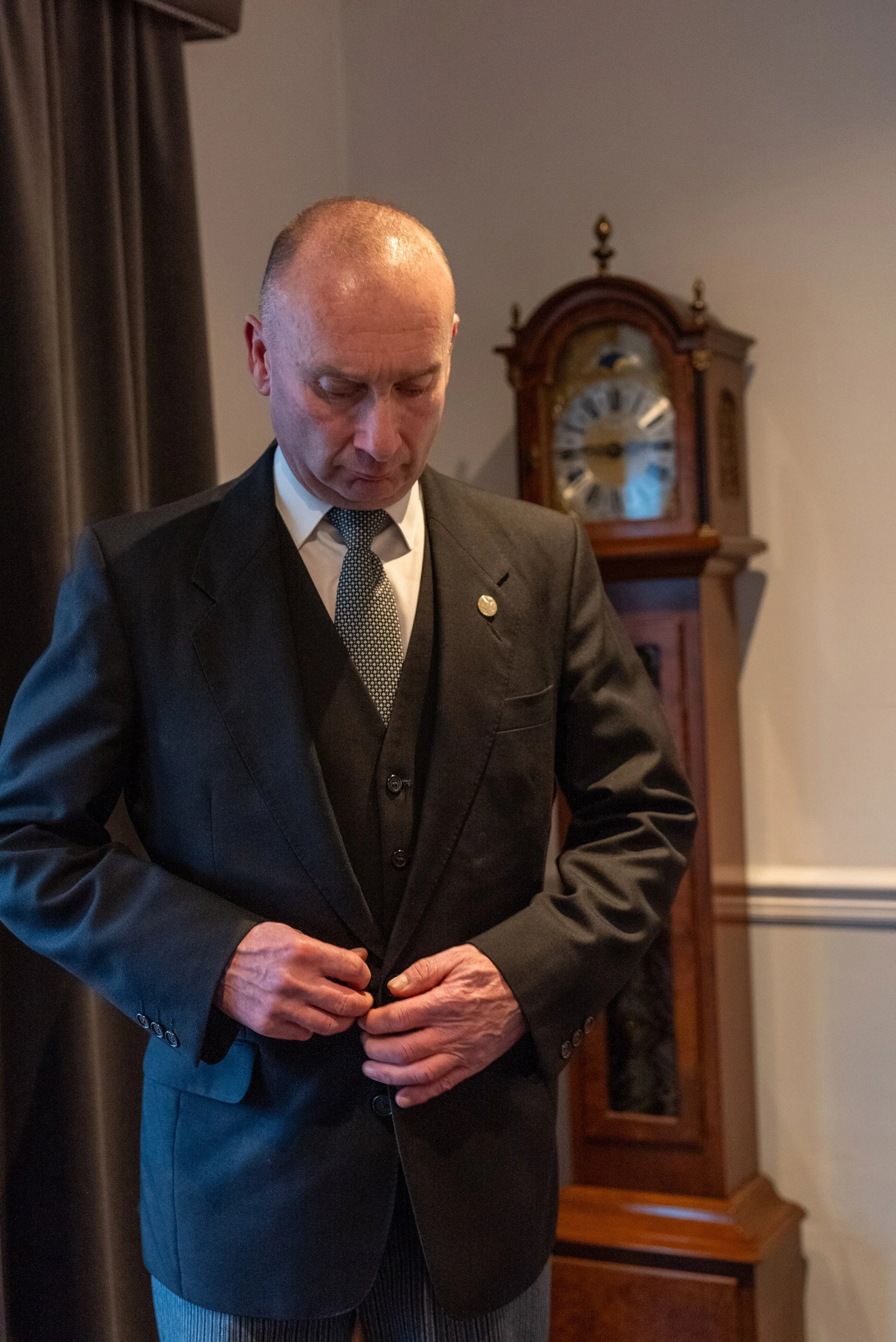 A man in a formal black suit adjusting his jacket in a room with a grandfather clock in the background.